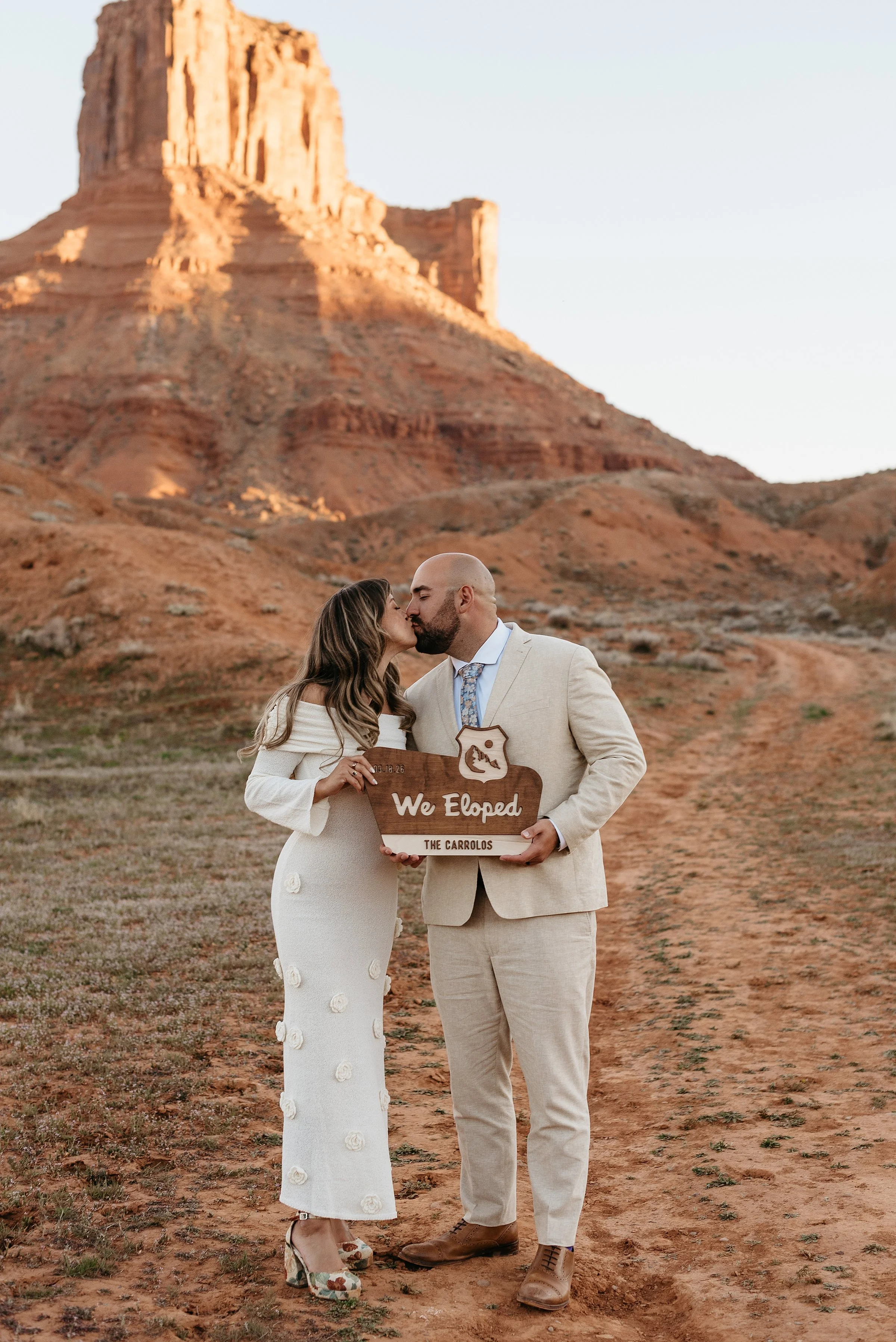A bride and groom are kissing while holding a sign that says we eloped. They are standing in a red rock desert with a large butte in the background behind them.