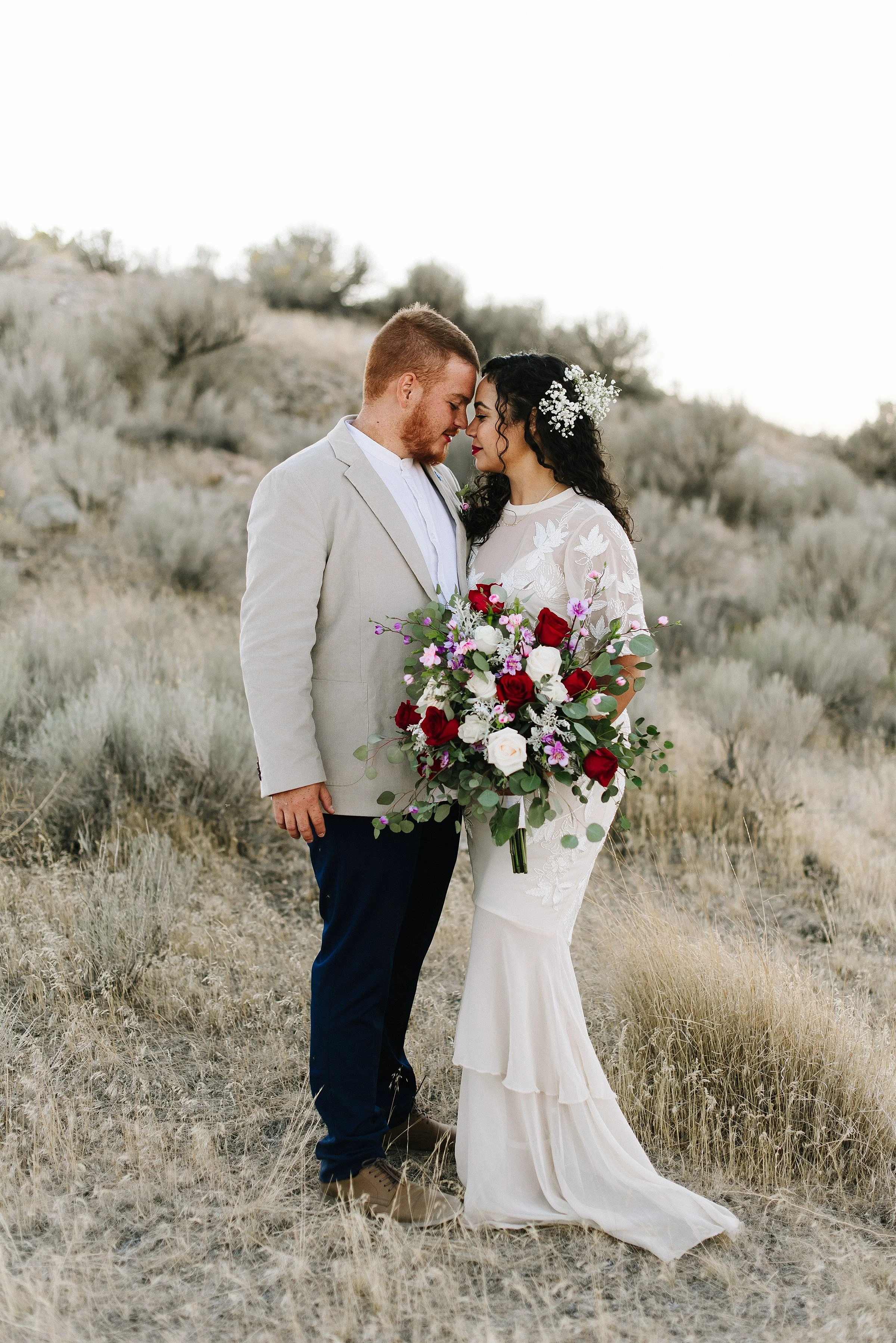 A bride and groom are embracing while holding a large bouquet. They are standing in a sage desert.