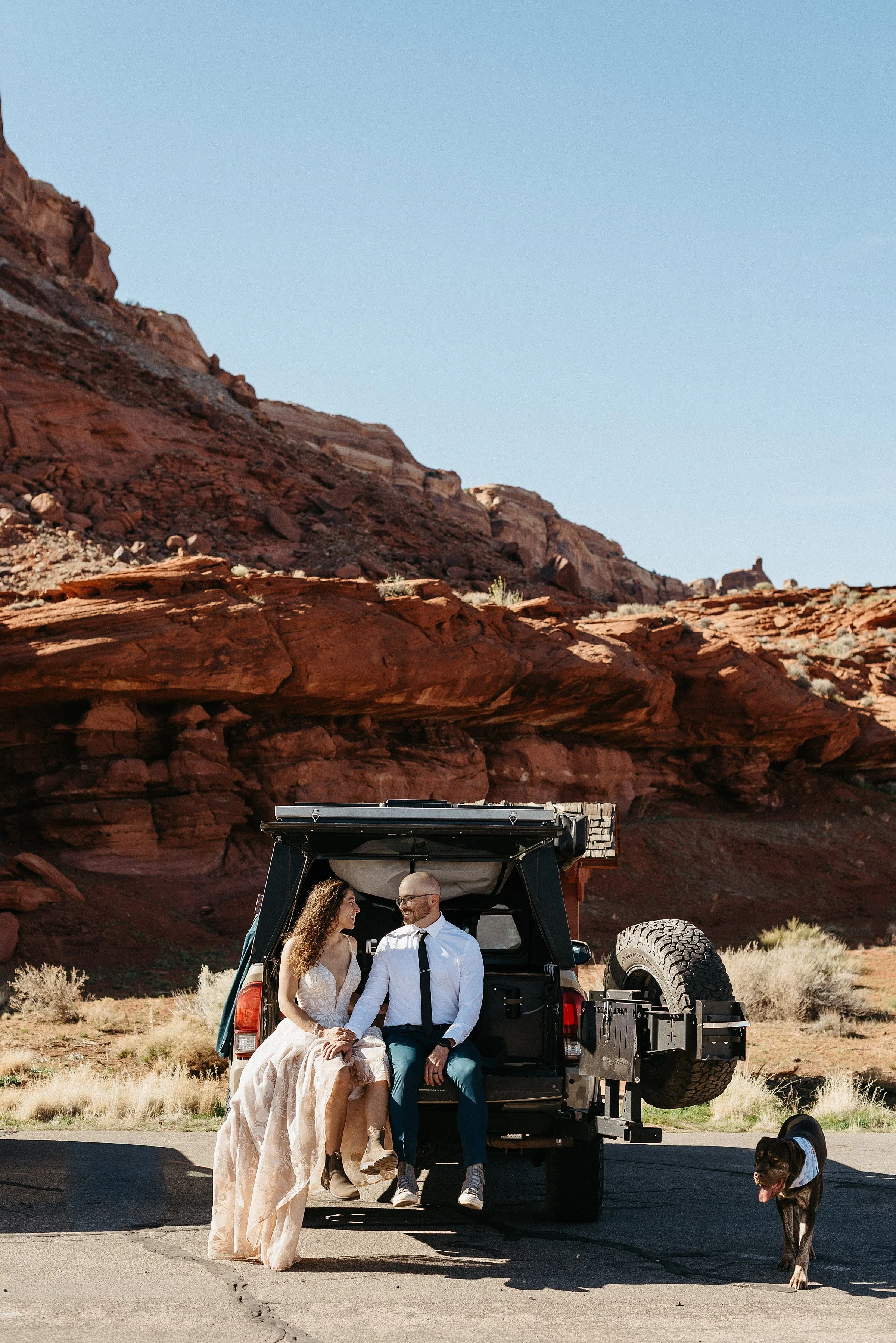 A bride and groom are sitting in the back of a Toyota Tacoma in a red rock desert. They are looking at each other.