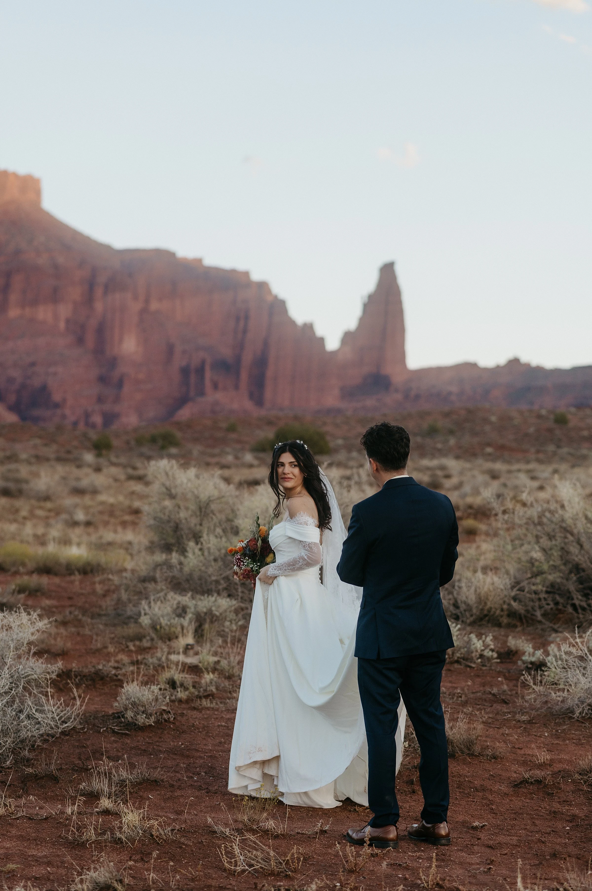 A bride and groom walk into a red rock desert. The bride is looking back at the groom and he is holding the train of her dress. There is a tall red rock wall formation in the background, and light green sage and plants around them.