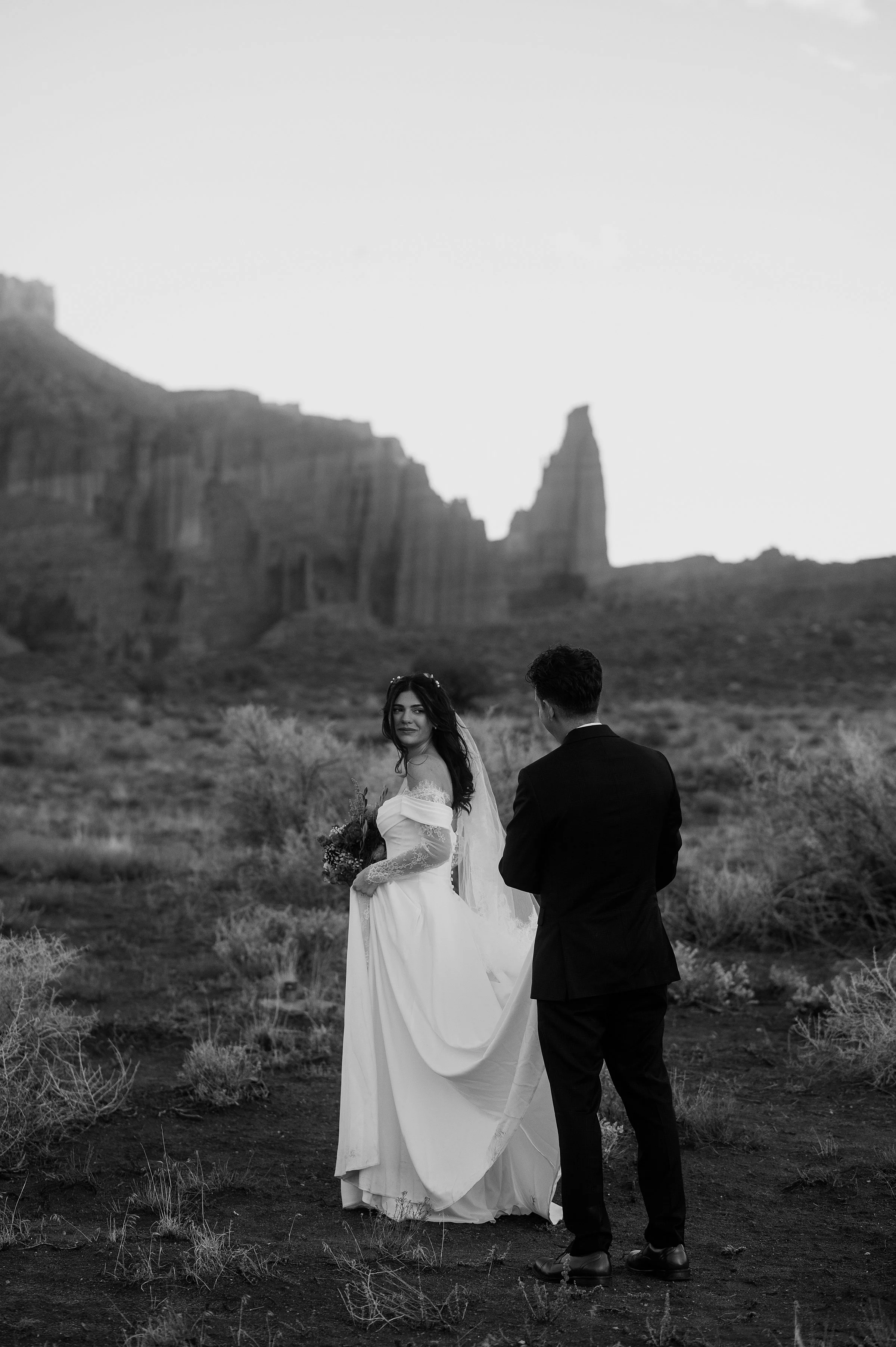 A bride and groom are walking in the desert. The bride is holding her dress and looking back over her shoulder at the groom. The photo is black and white and there are jagged rock spires in the background.
