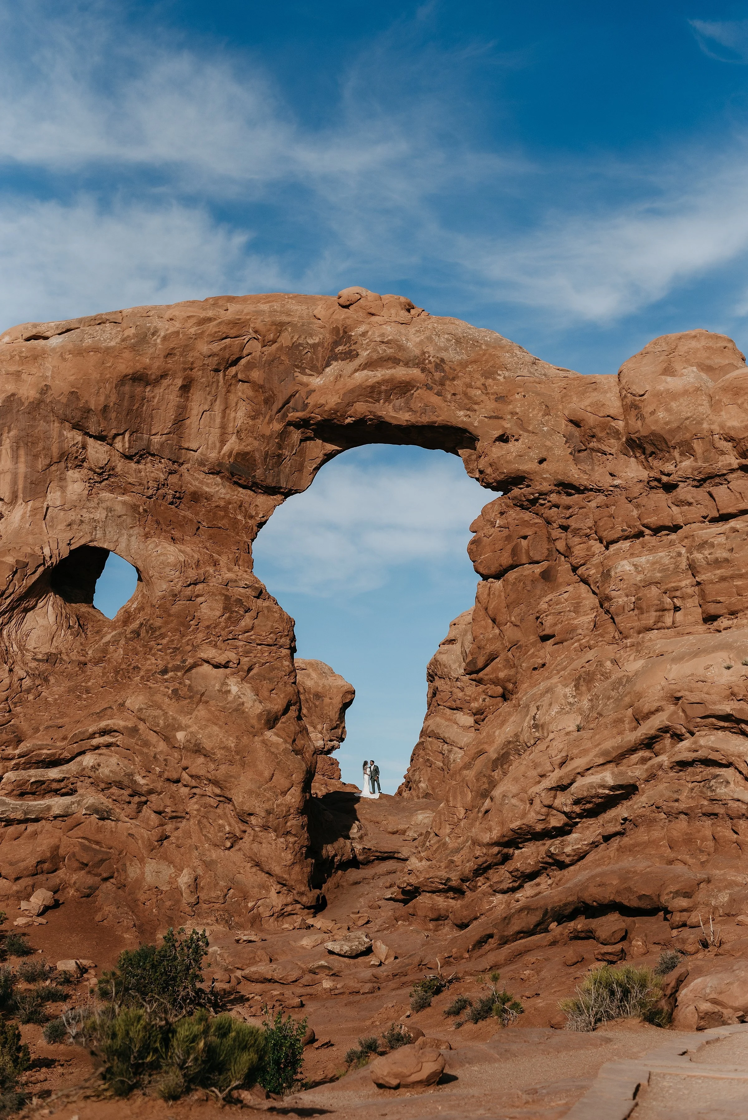 A bride and groom are embracing in a large red rock arch. The sky is blue behind them and there is greenery in the foreground.
