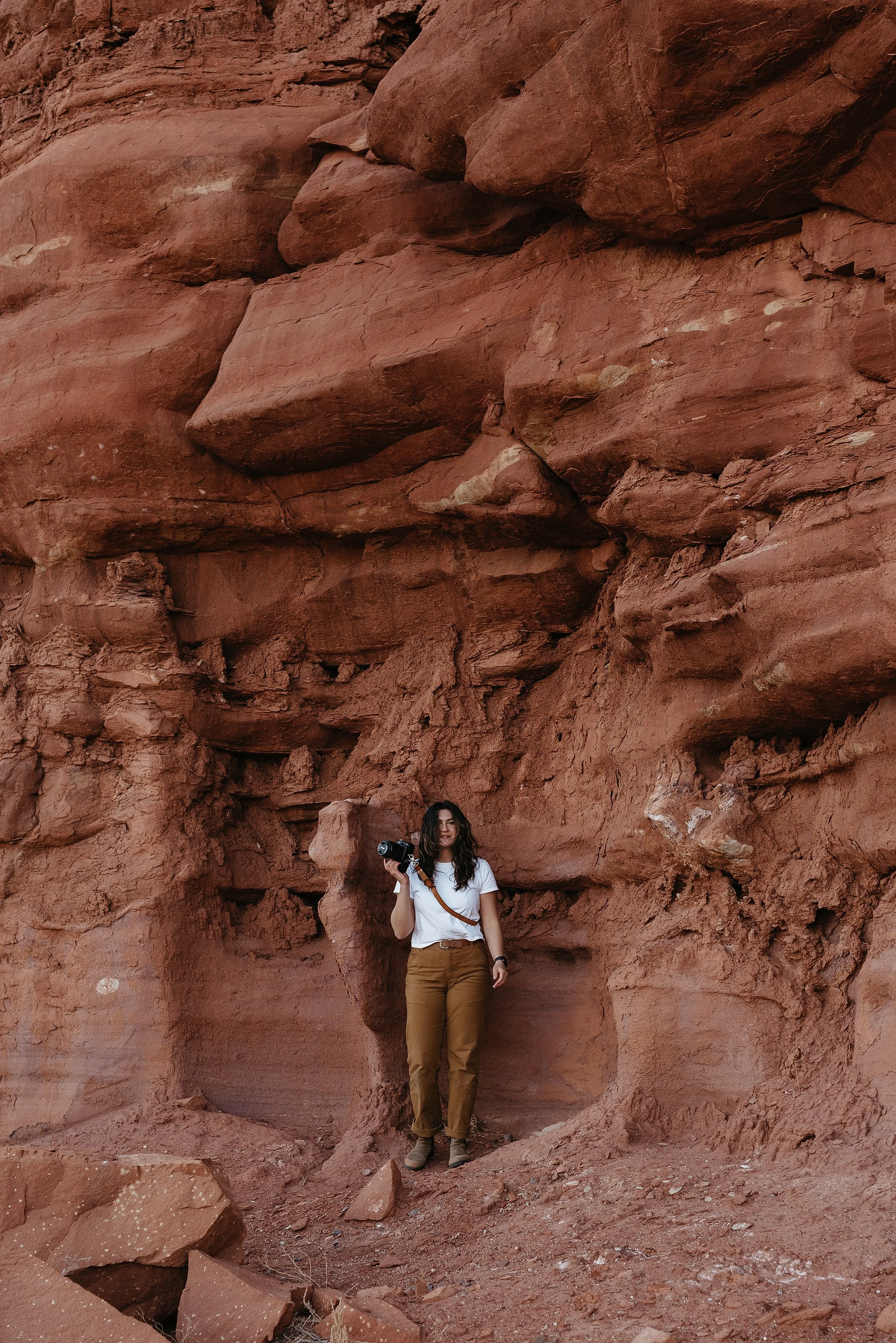 A woman is wearing a white t shirt and carhartt pants. she is holding a camera in her right hand and standing against a red rock wall.