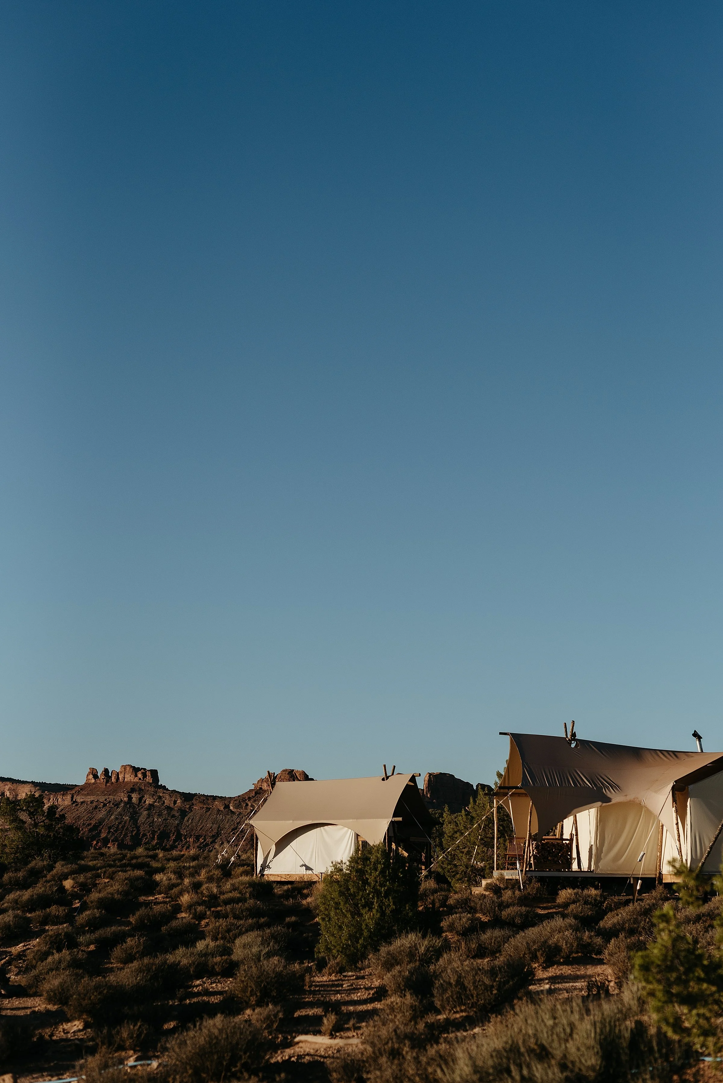Two canvas tents are in a desert landscape with red rock cliffs in the background. the sun is rising and the sky is very blue.