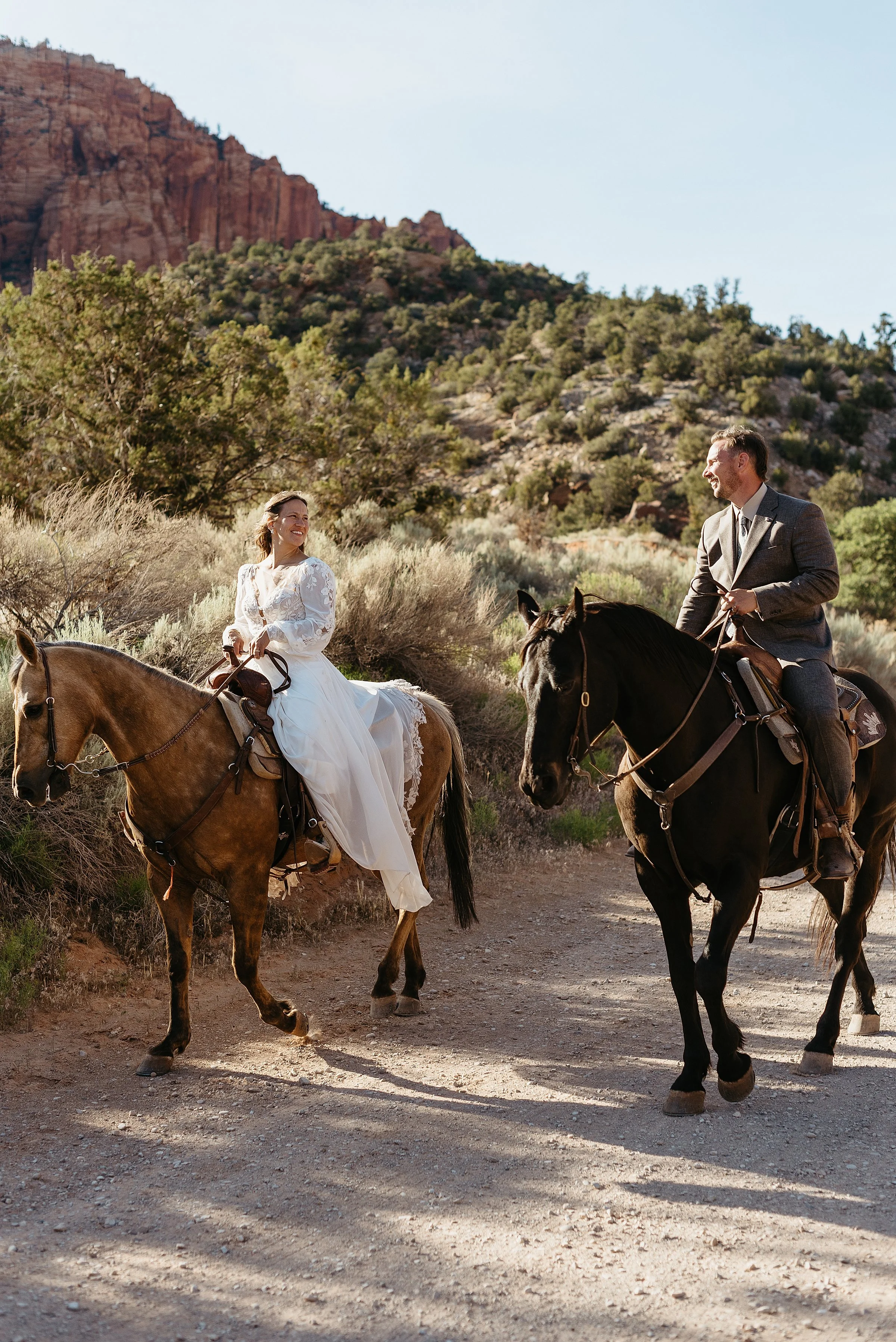 A bride and groom are riding horses in a red rock desert