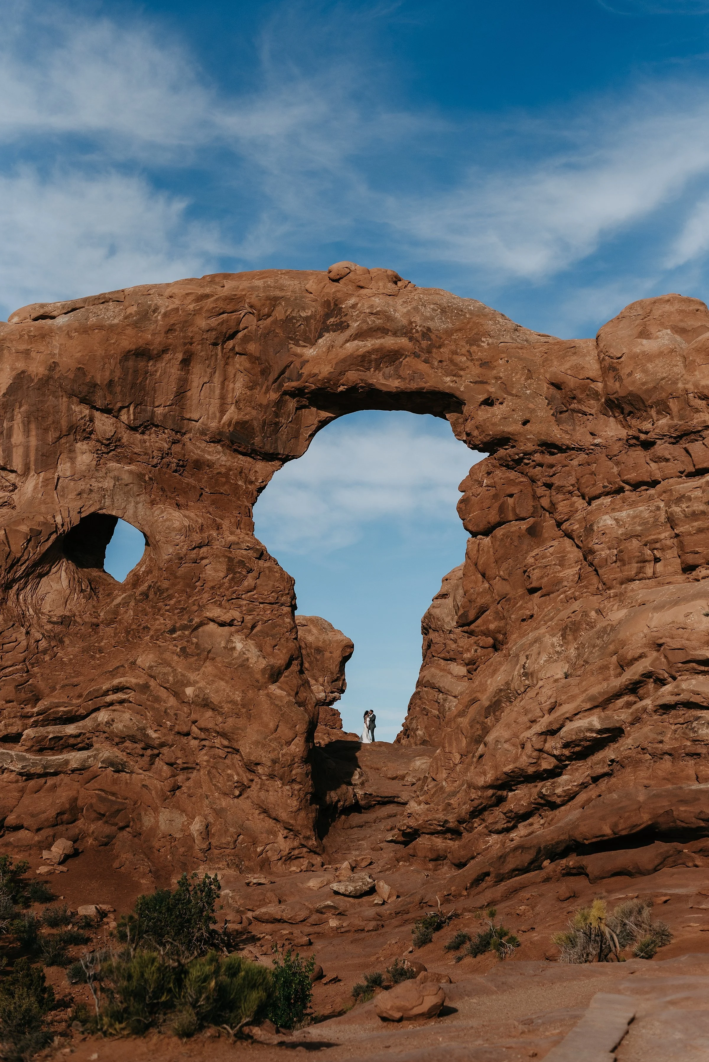 A bride and groom are embracing in a large red rock arch in arches national park. The blue sky has wispy clouds and is visible through the rock arch and a smaller round arch on the left. There are some green desert plants in the foreground.
