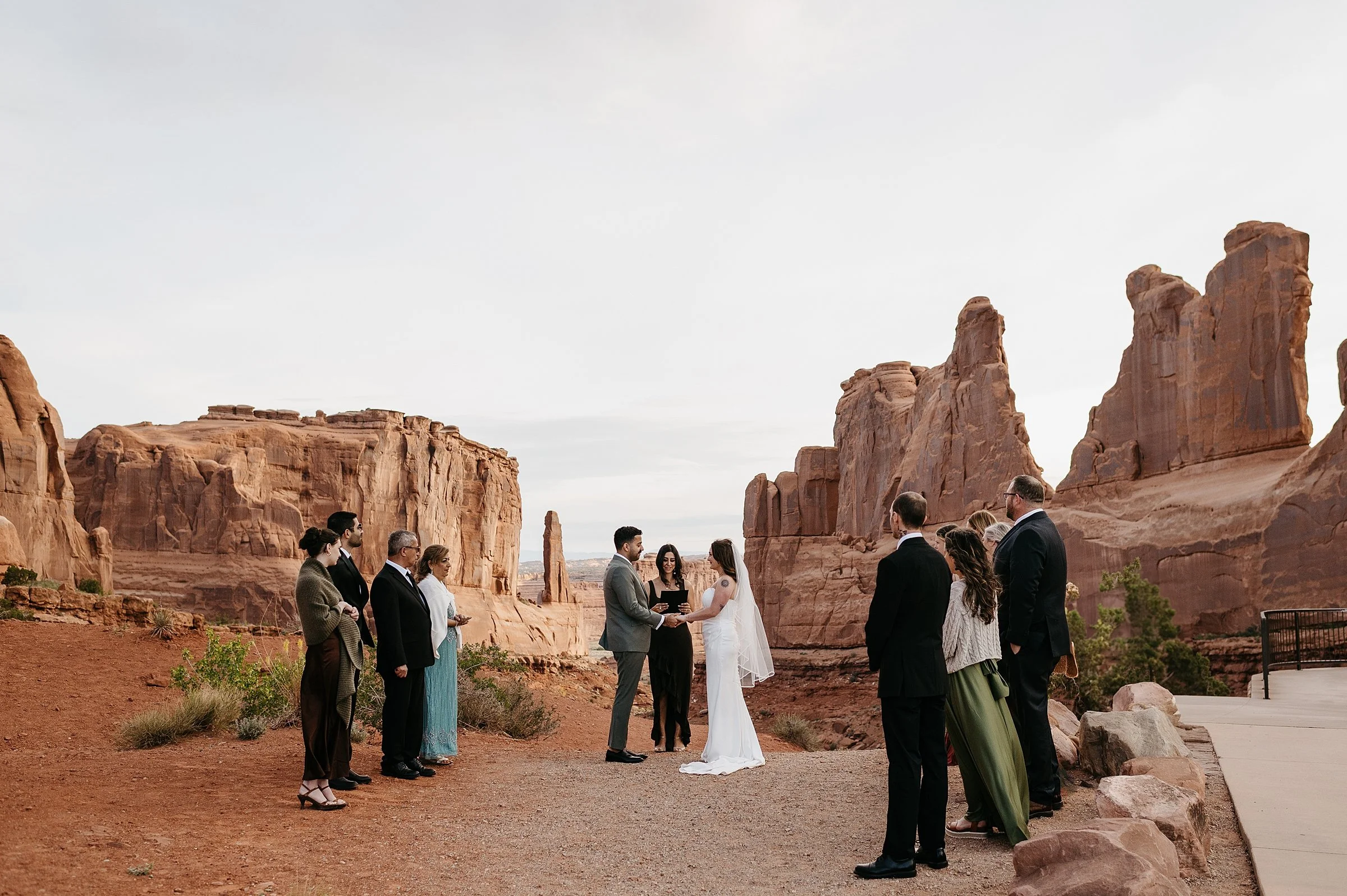 A bride and groom stand in Arches National Park during their wedding ceremony hand in hand. The couple has friends and family on either side witnessing their ceremony up against large red rock walls.