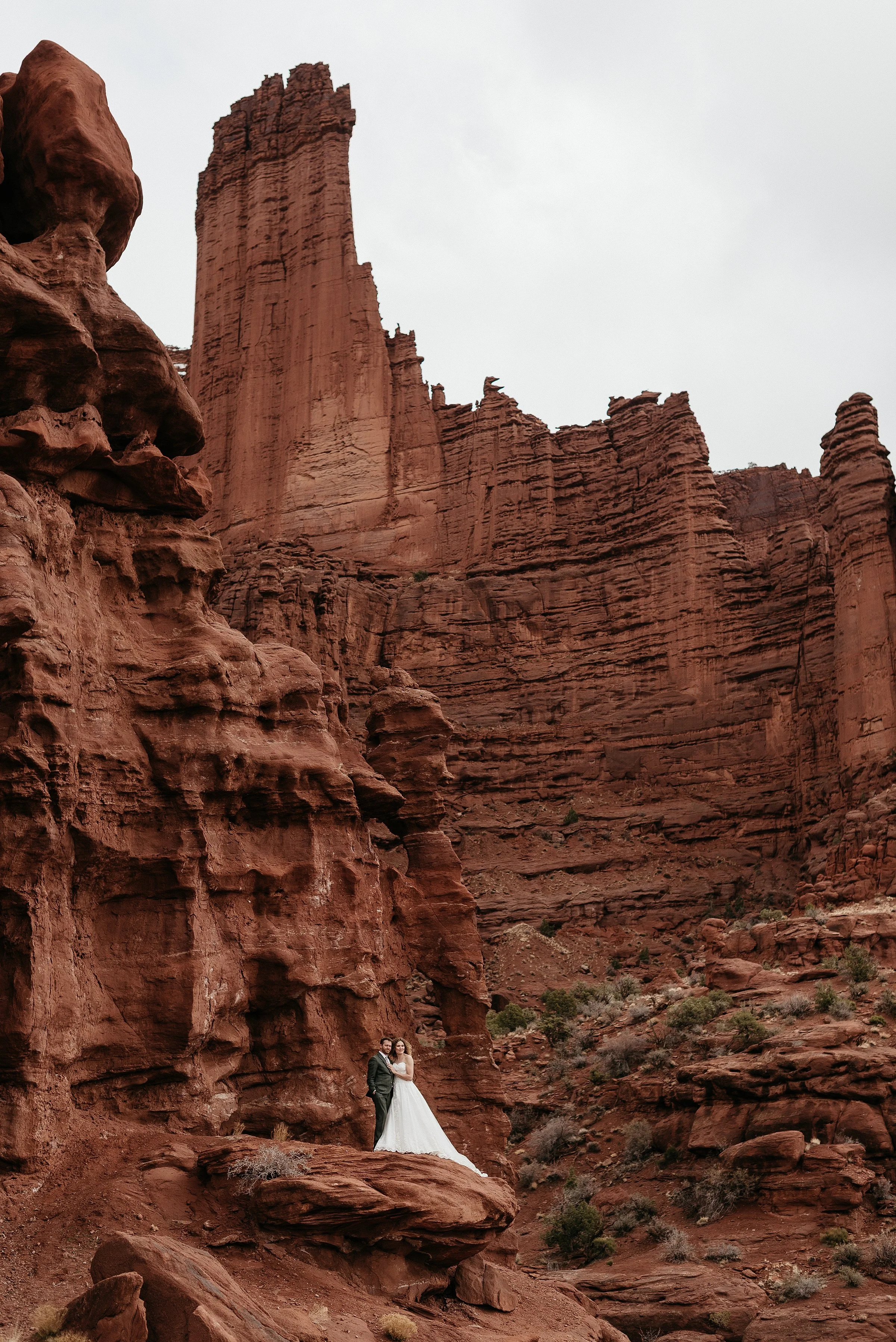 A bride and groom are standing in front of a large red rock wall in the desert. They are embracing and looking off to the right of the image.
