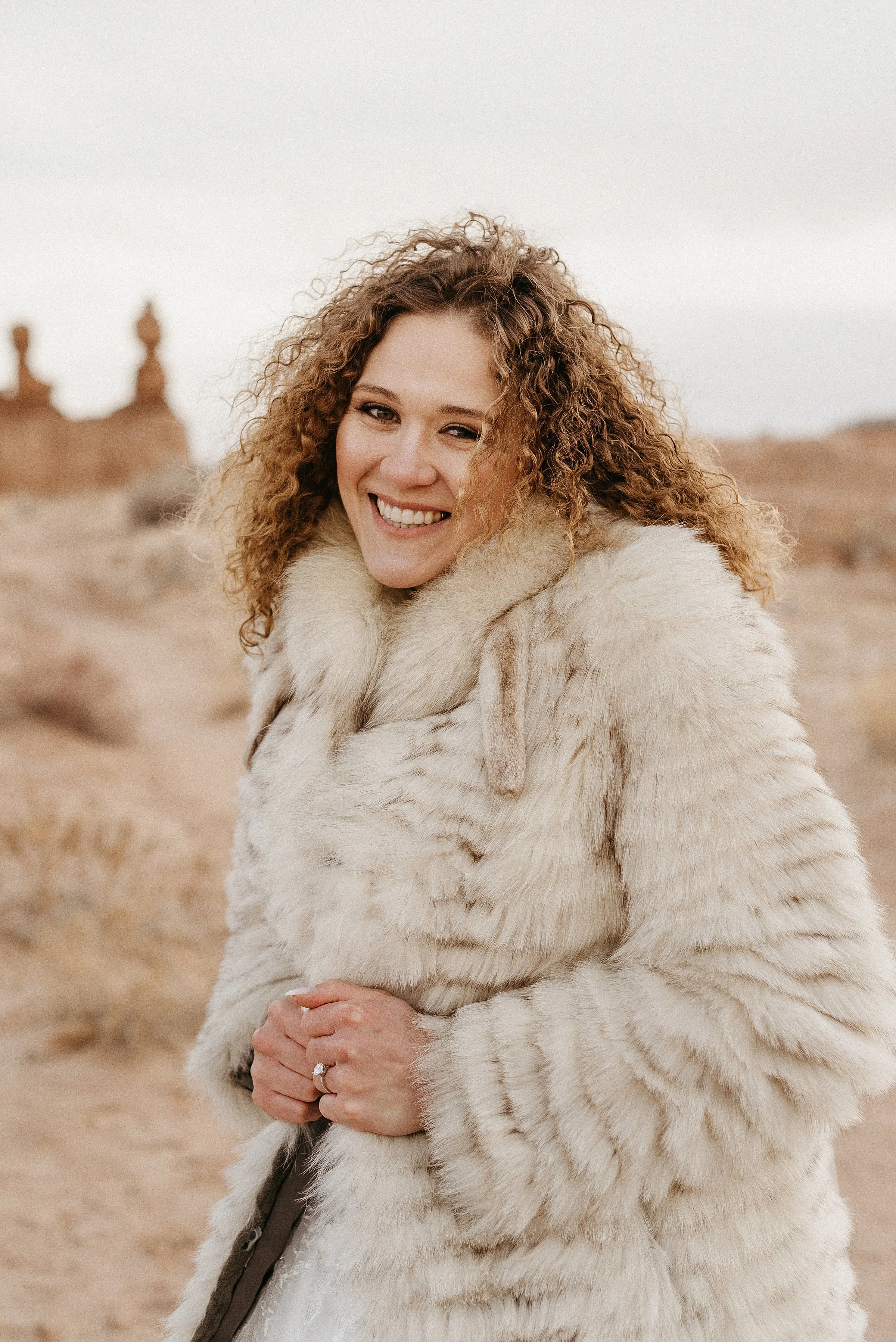 A bride is wearing a vintage fur coat over her wedding dress in a red rock desert. She has curly hair and a big smile.