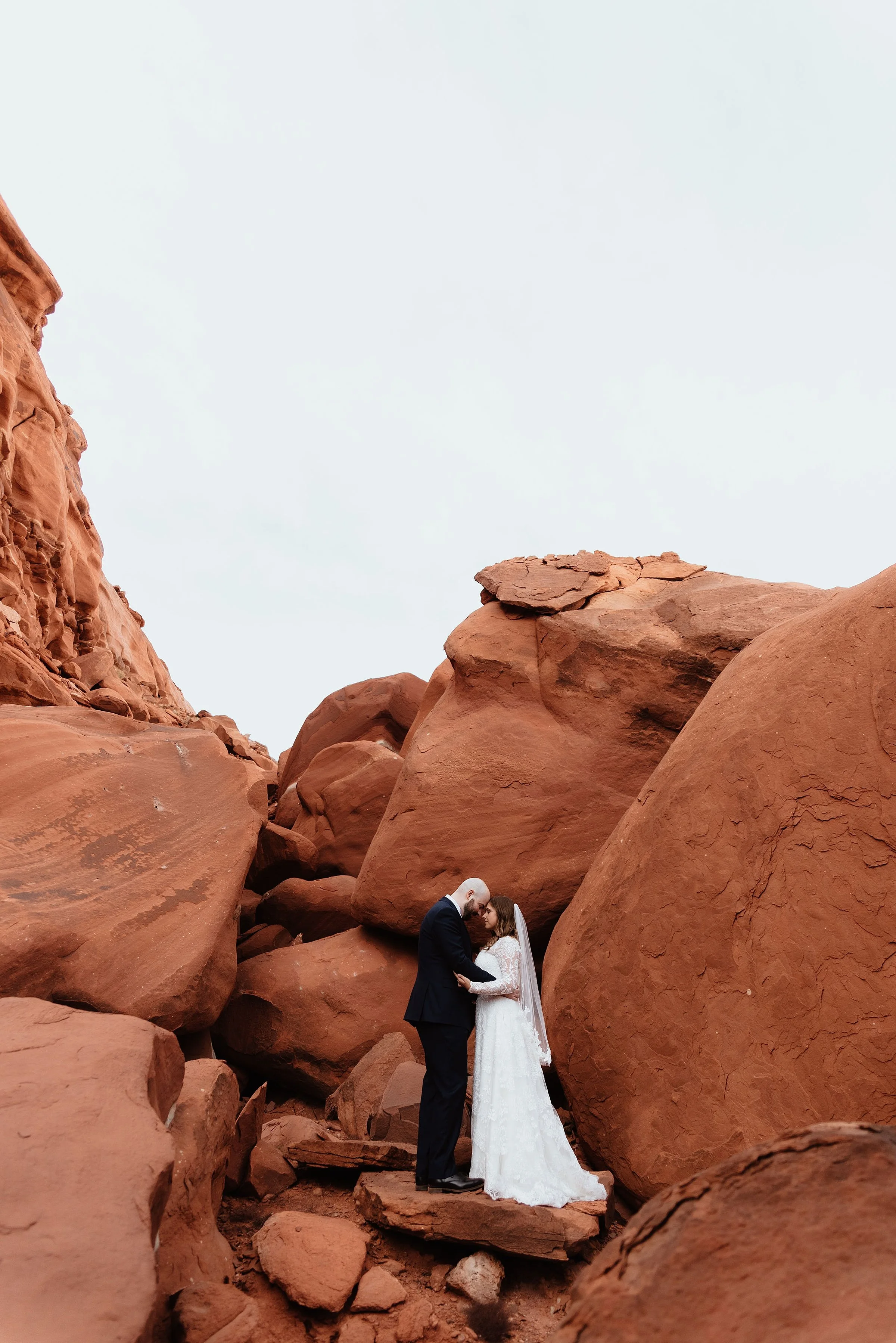 A bride and groom are embracing in the middle of a red rock boulder field.