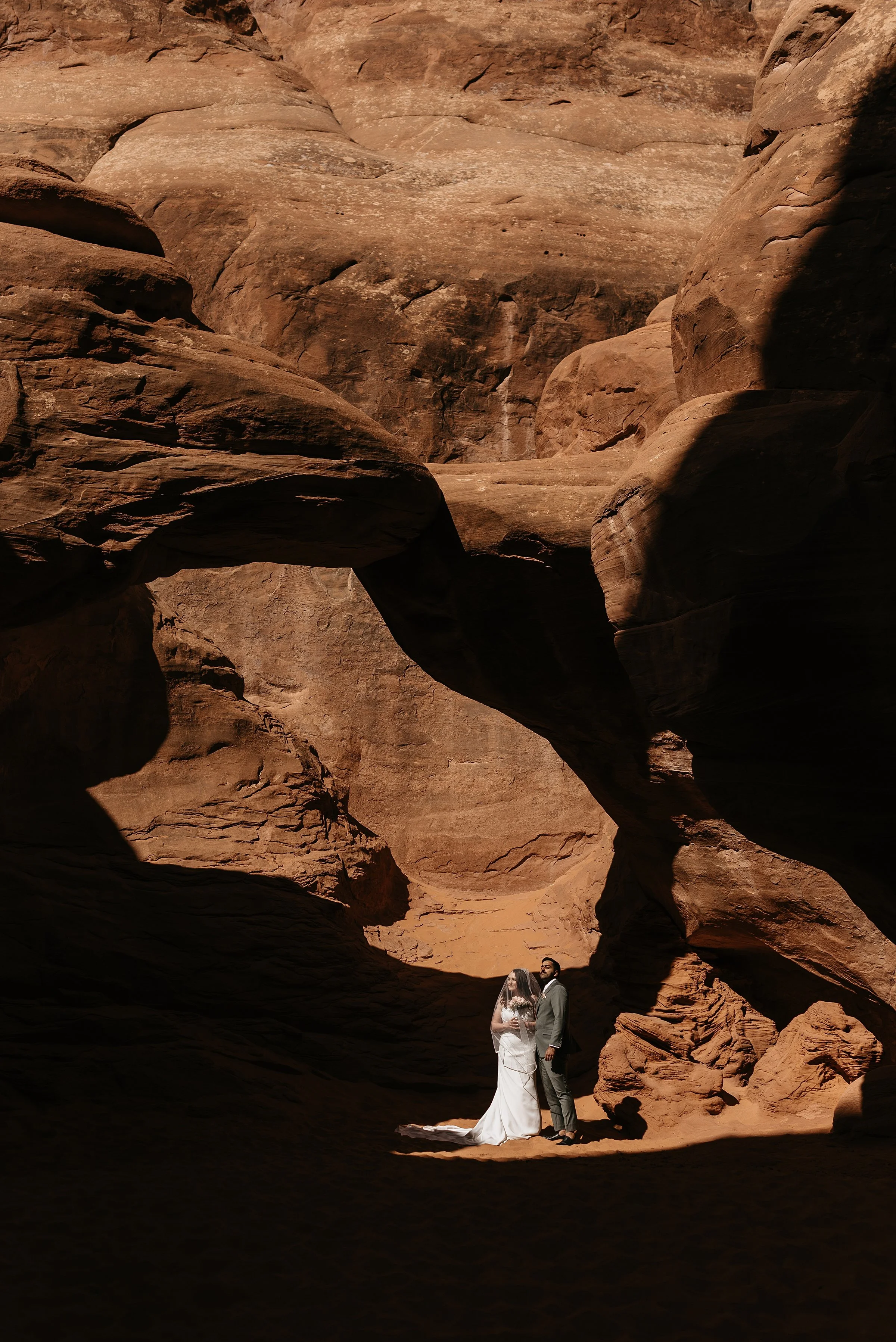 A bride and groom stand in the sun underneath Sand Dune Arch in Arches National Park. The bride's veil is over her face and she holds pink roses. The groom is wearing a sage green suit.