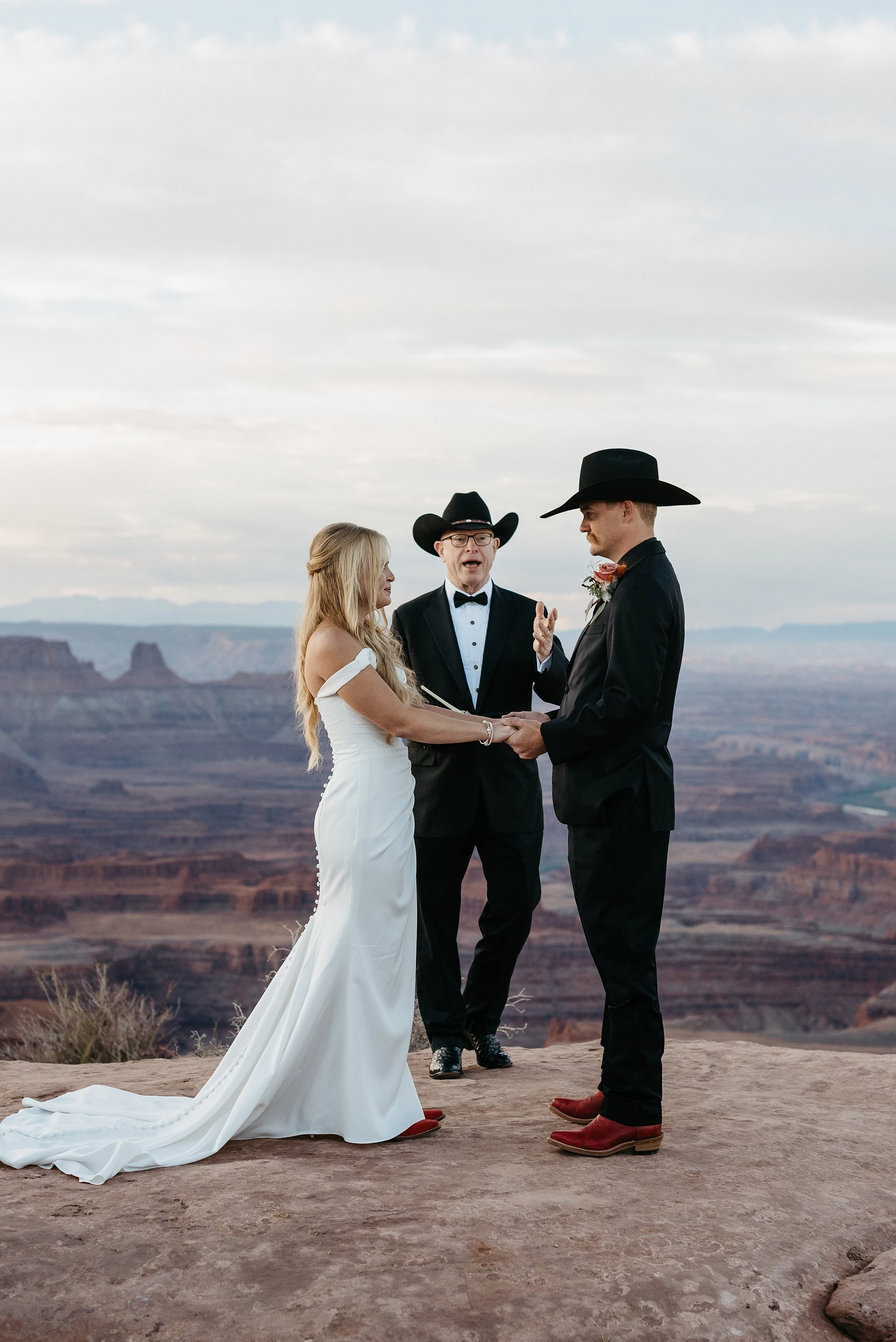 A bride and groom are facing each other holding hands during their wedding ceremony. A man in a tux and a cowboy hat is standing behind them officiating. They are on the edge of a cliff and there is a vast red rock canyon in the distance.
