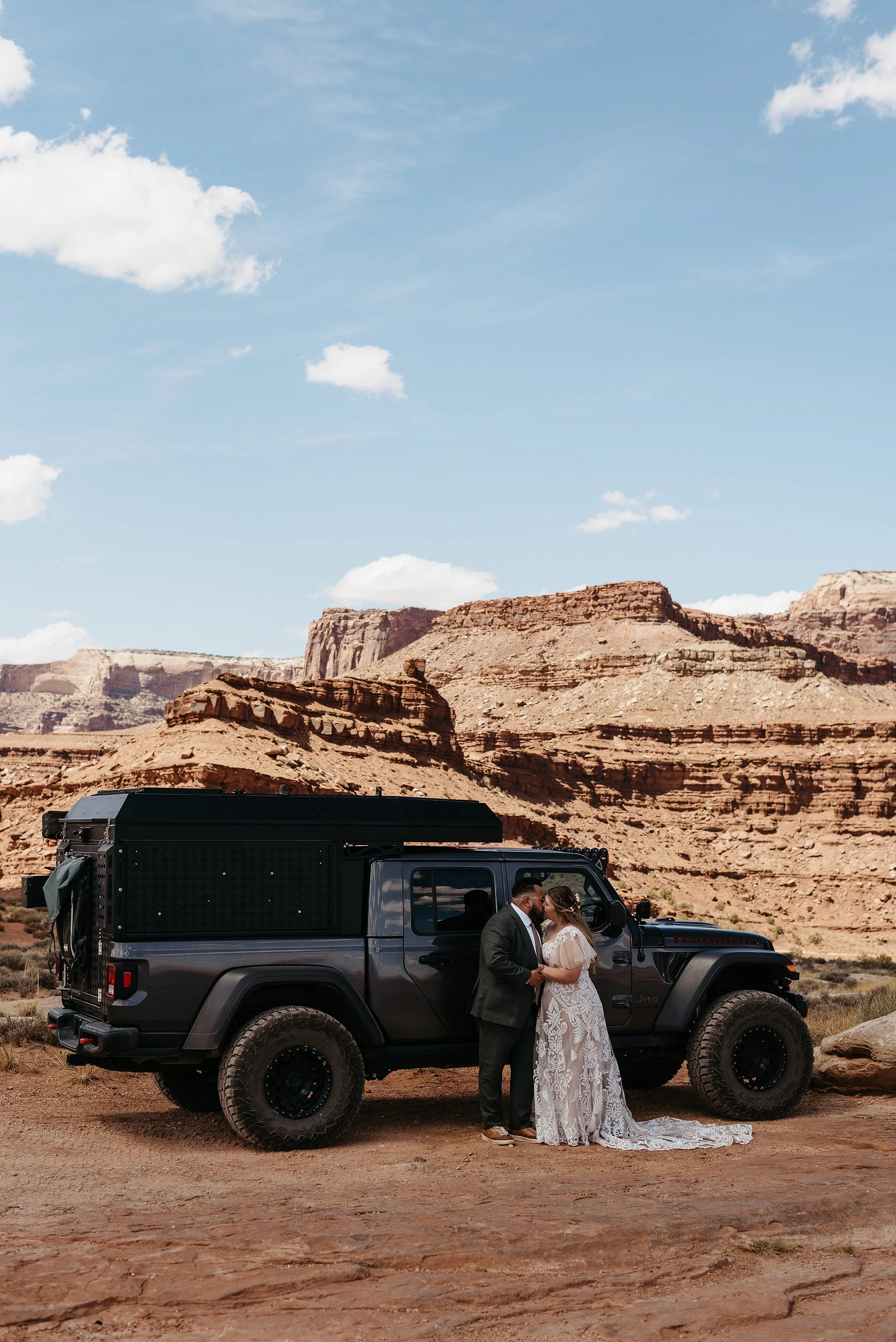 A bride and groom are kissing in front of their heavily decked out Jeep truck. The bride has a long train and there is a large red rock butte behind them.