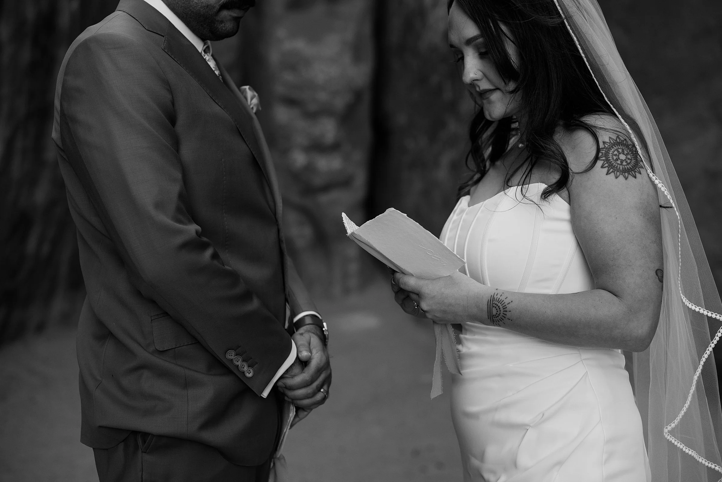 A bride and groom are facing each other and the bride is holding a vow book. She has a nose piercing and a tattoo of a rising sun on her left wrist.