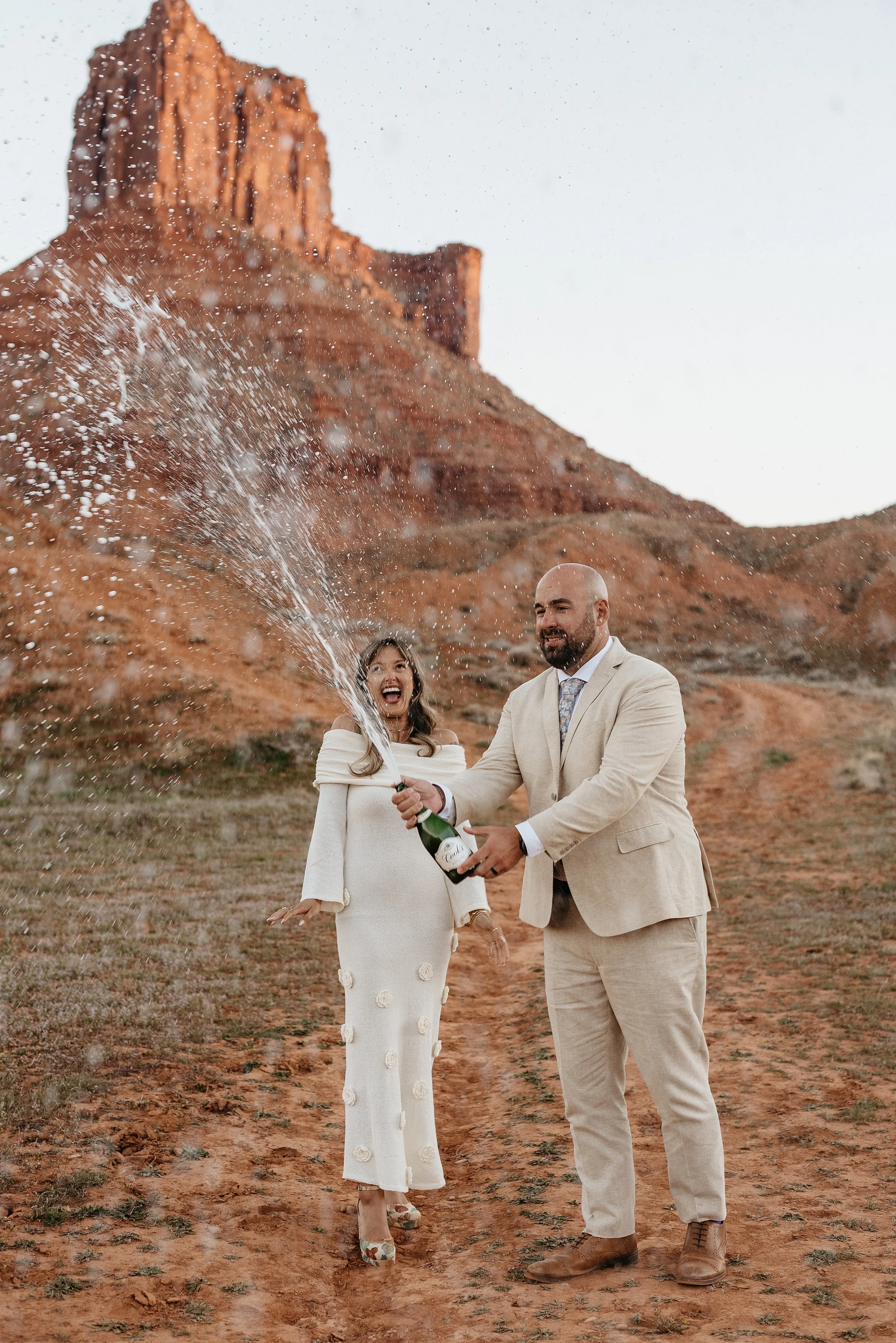 A bride and groom are standing in a red rock desert. The groom is opening a bottle of champagne that is spraying everywhere.