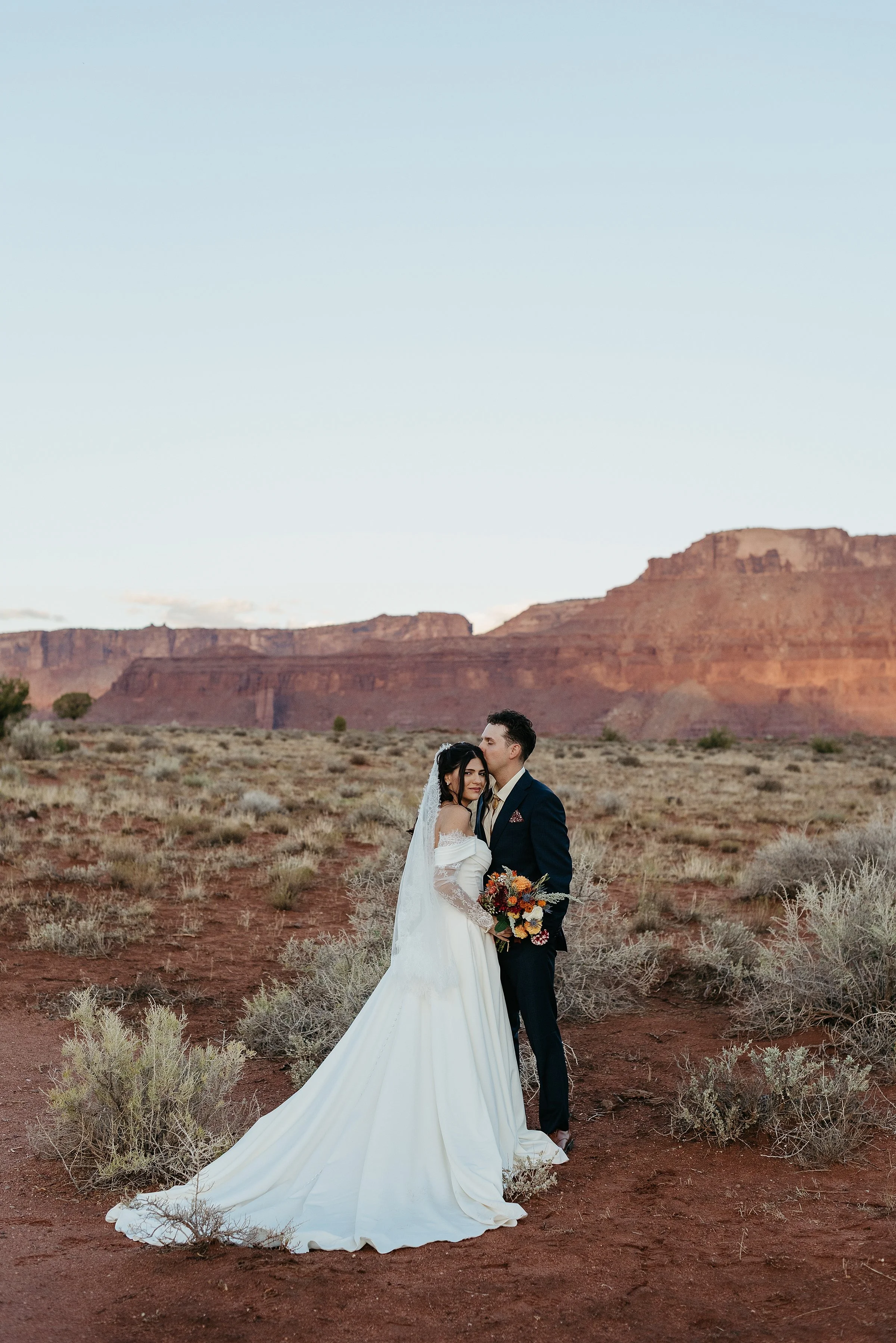 A bride and groom stand together in a red rock desert. the groom is kissing the brides head and she is smiling at the camera holding a bouquet of wildflowers. the sun is setting and the red rock walls in the background are shades of purple and orange