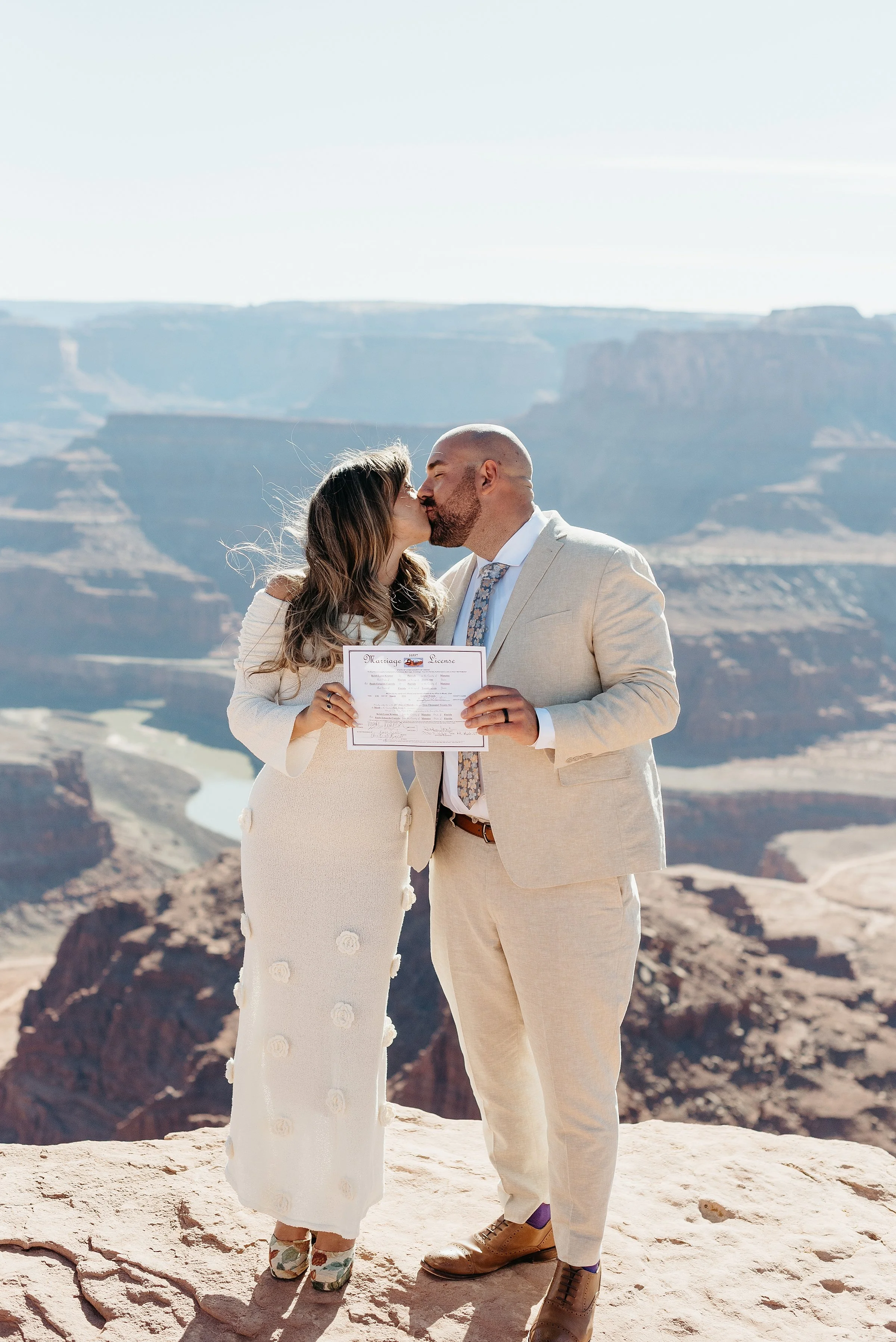 A bride and groom stand on a desert cliff holding their marriage license. They are kissing.