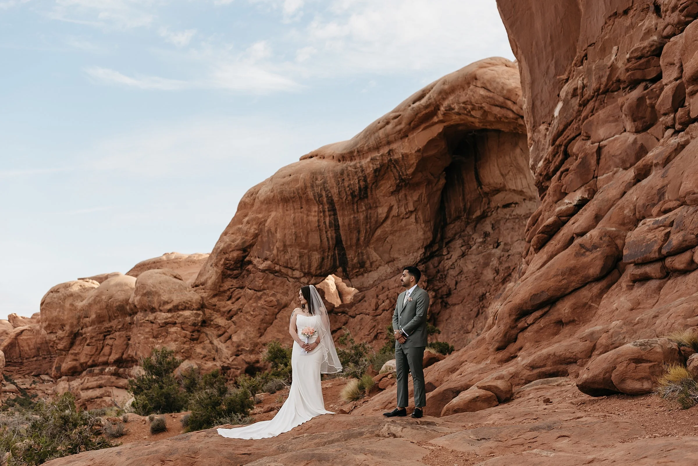 A bride and groom stand a few feet apart in a red rock desert. There is a small arch in the background between them. There is desert greenery around them and the couples is staring off to the left. The bride holds a bouquet of pink flowers.