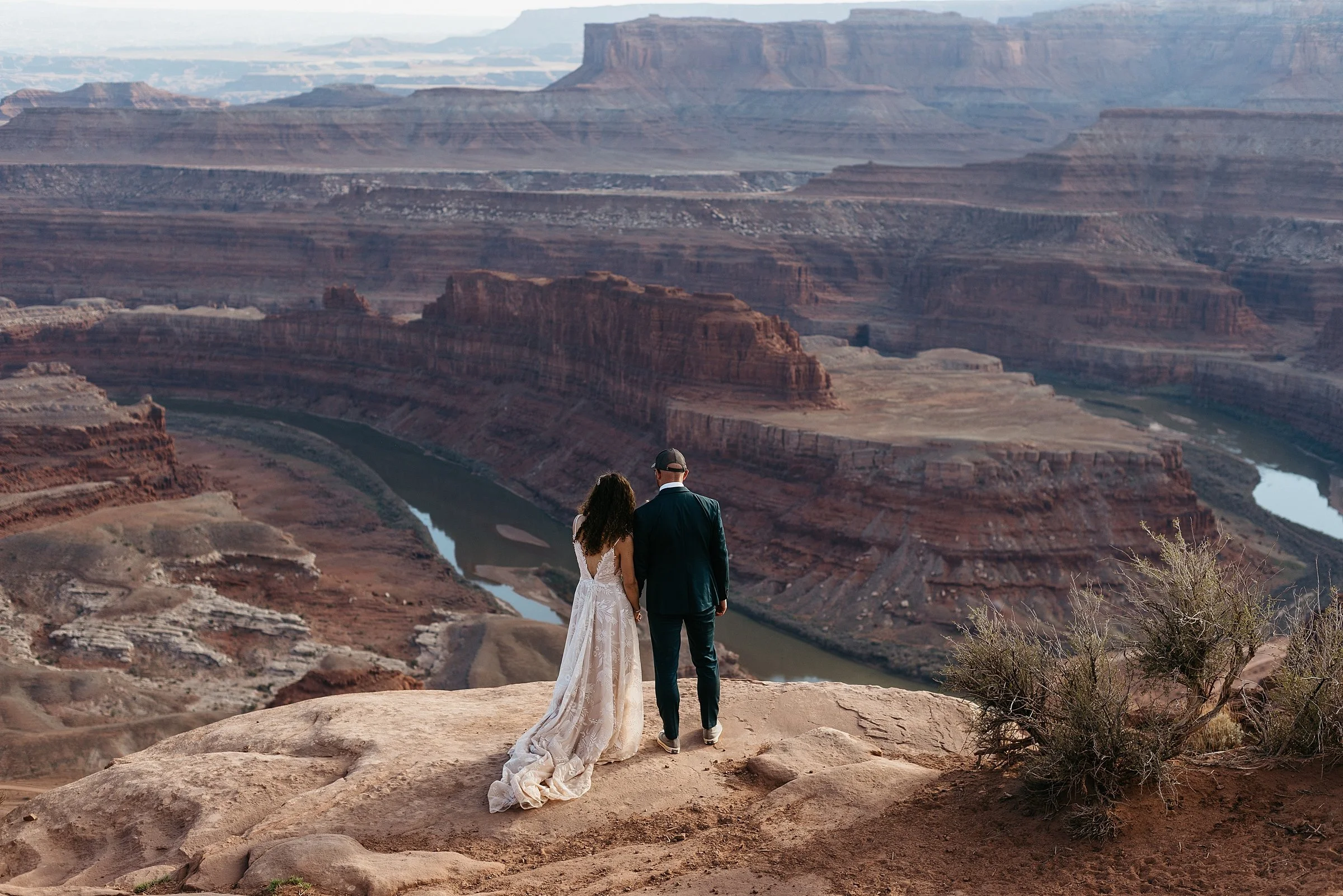 bride and groom are standing holding hands on a red rock cliff at Dead Horse Point overlooking a desert canyon with a river flowing through it. The sun is setting and the canyon is various shades of red, purple, and orange.