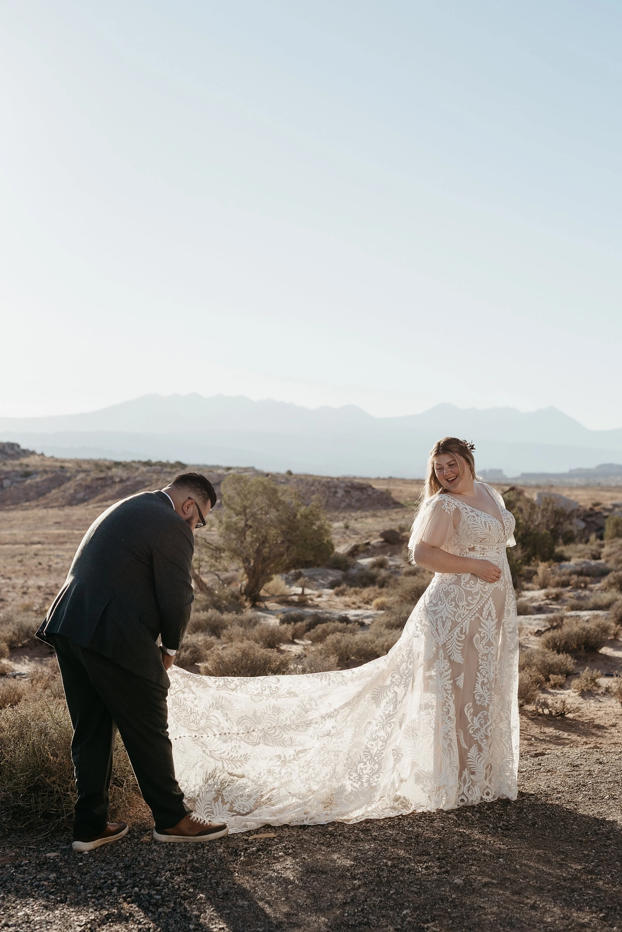 A groom is adjusting a bride's lace wedding dress train. The sun is hitting them from behind and there is a mountain range in the background across a open desert area.