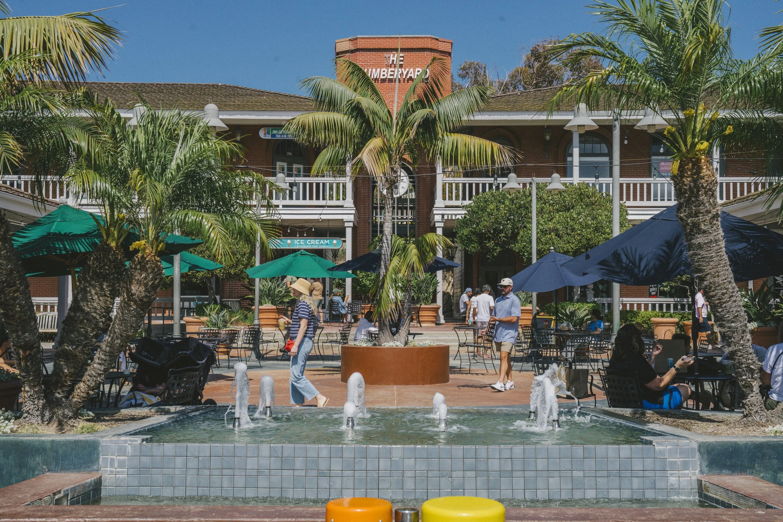 A lively outdoor plaza with palm trees, umbrellas, and people sitting and walking around, with a red brick building in the background and a small fountain in the foreground.