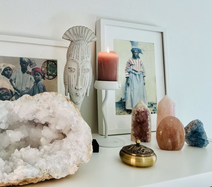Altaar voor helderziende reading en energy healing met decoratieve items en foto's op een witte tafel, waaronder een grote kristallen rots, kaarsen, een foto van een vrouw in traditionele surinaamse kleding, en beelden van mensen en een masker.