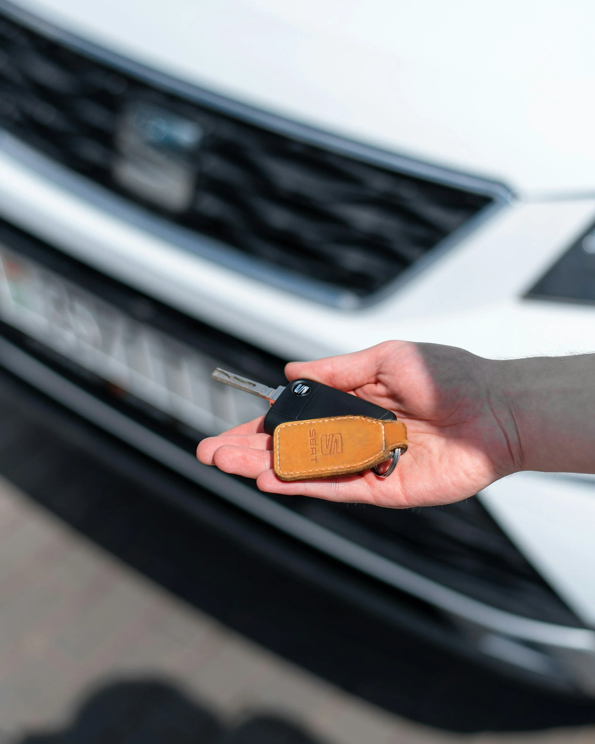 A person's hand holding car keys with a brown leather keychain in front of a white car's front grille.
