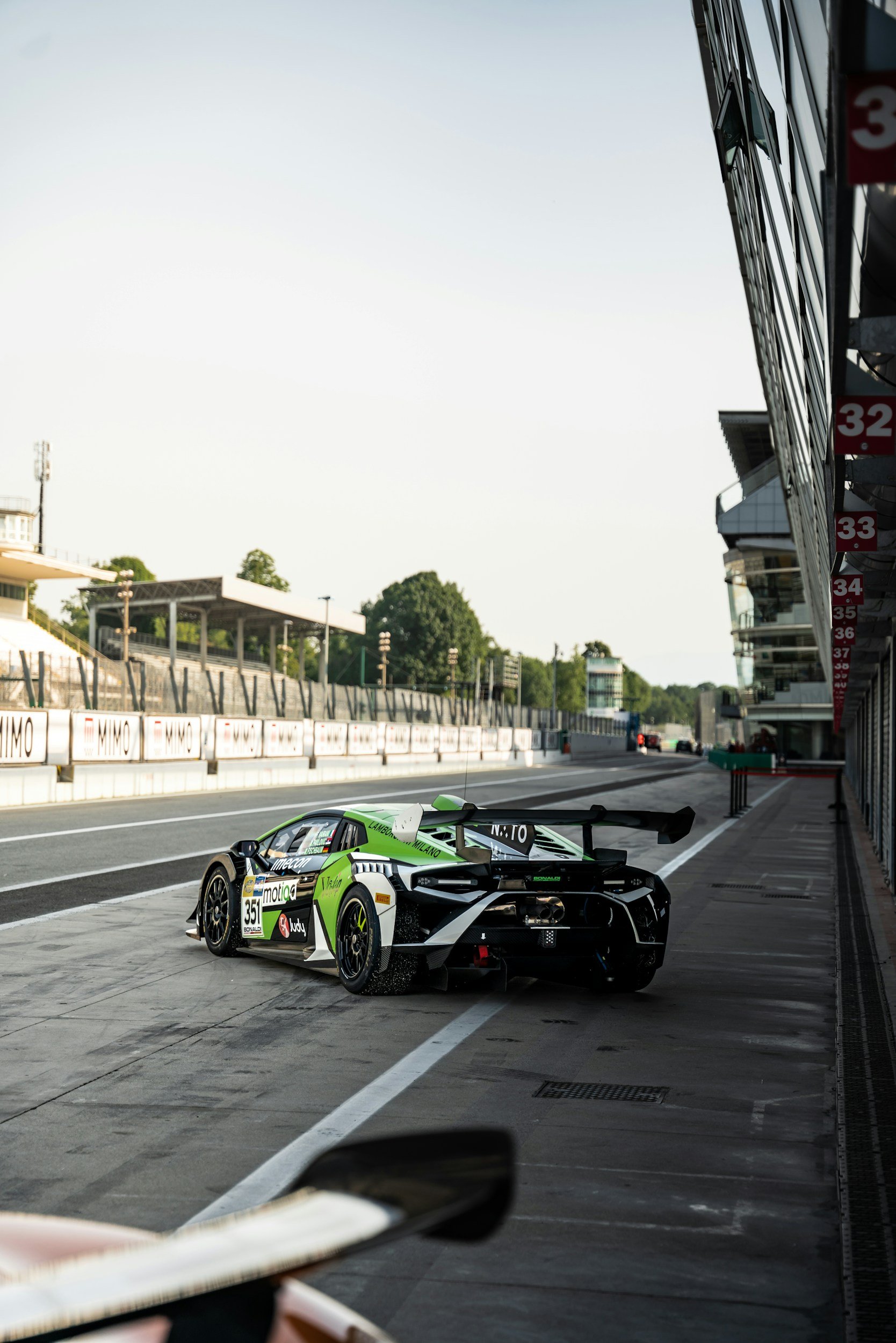 A green and black race car in the pit lane of a racetrack with empty garage spaces to the right and a grandstand in the background.