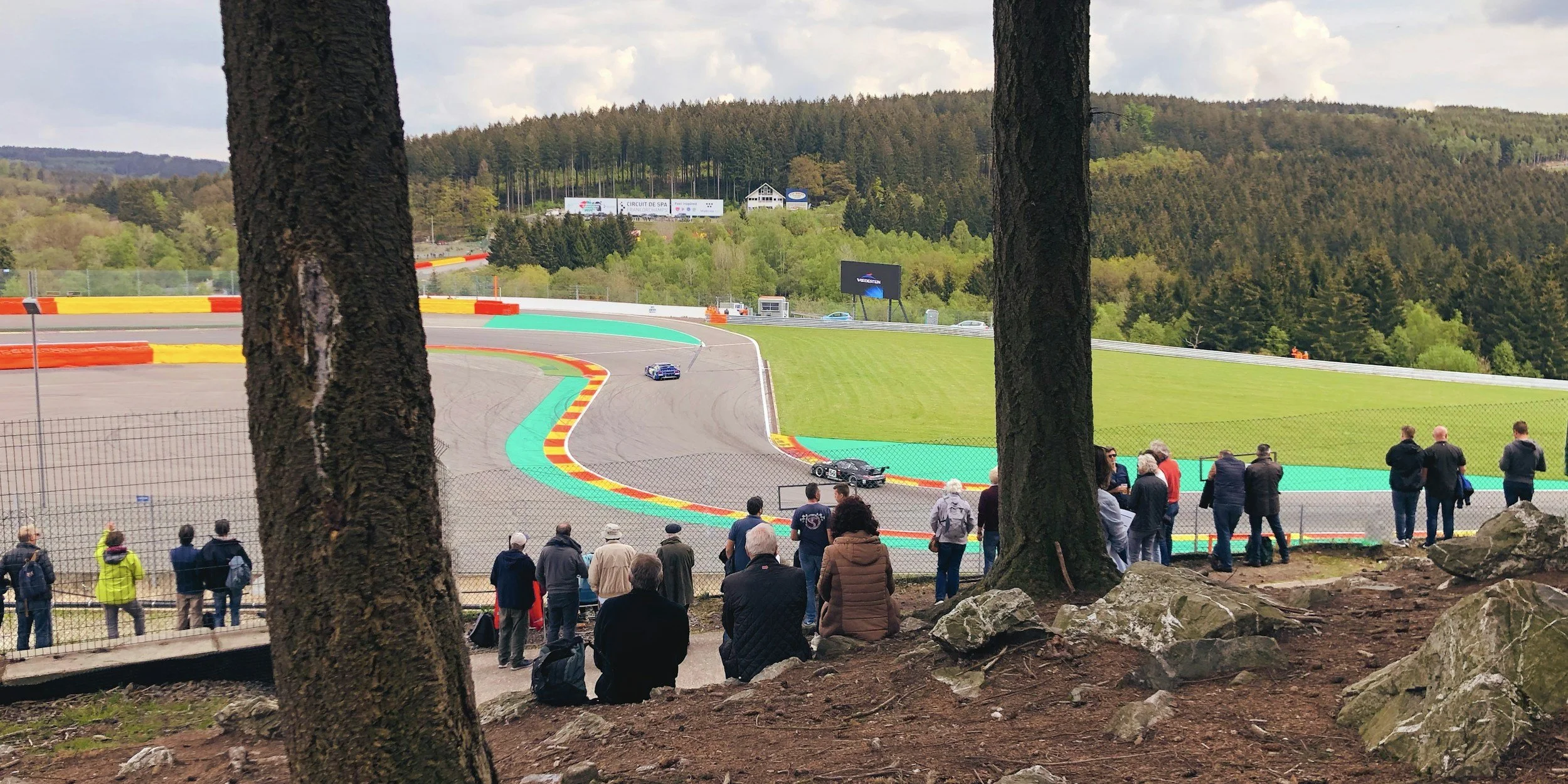 View of a race track with colorful curbs and a racing car on the track, spectators watching from the hillside behind a fence, trees and forest in the background.