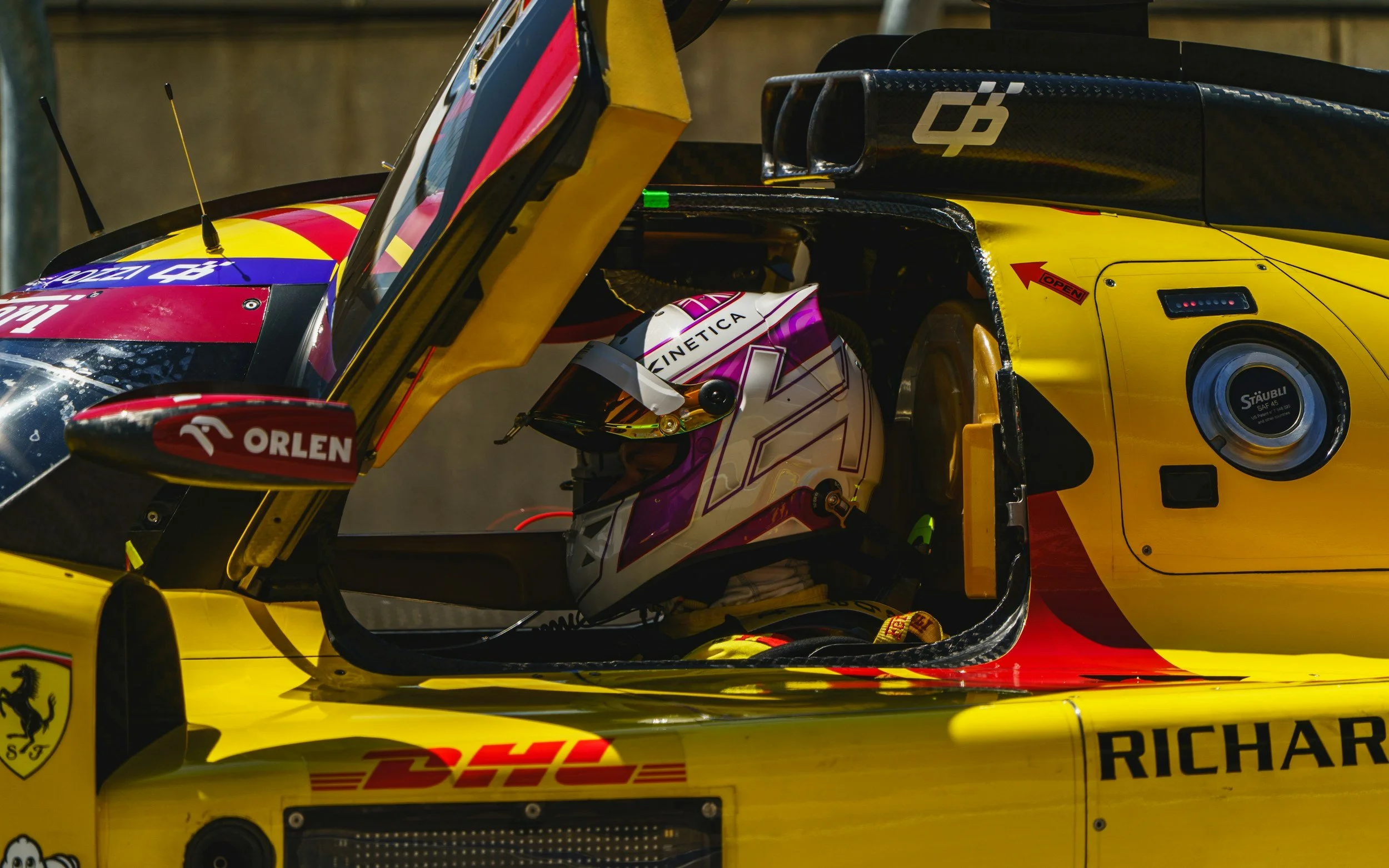 Close-up of a race car driver in yellow and red racing suit inside a yellow race car, wearing a purple and white helmet with the logo Kinetica, in a pit lane or garage area.
