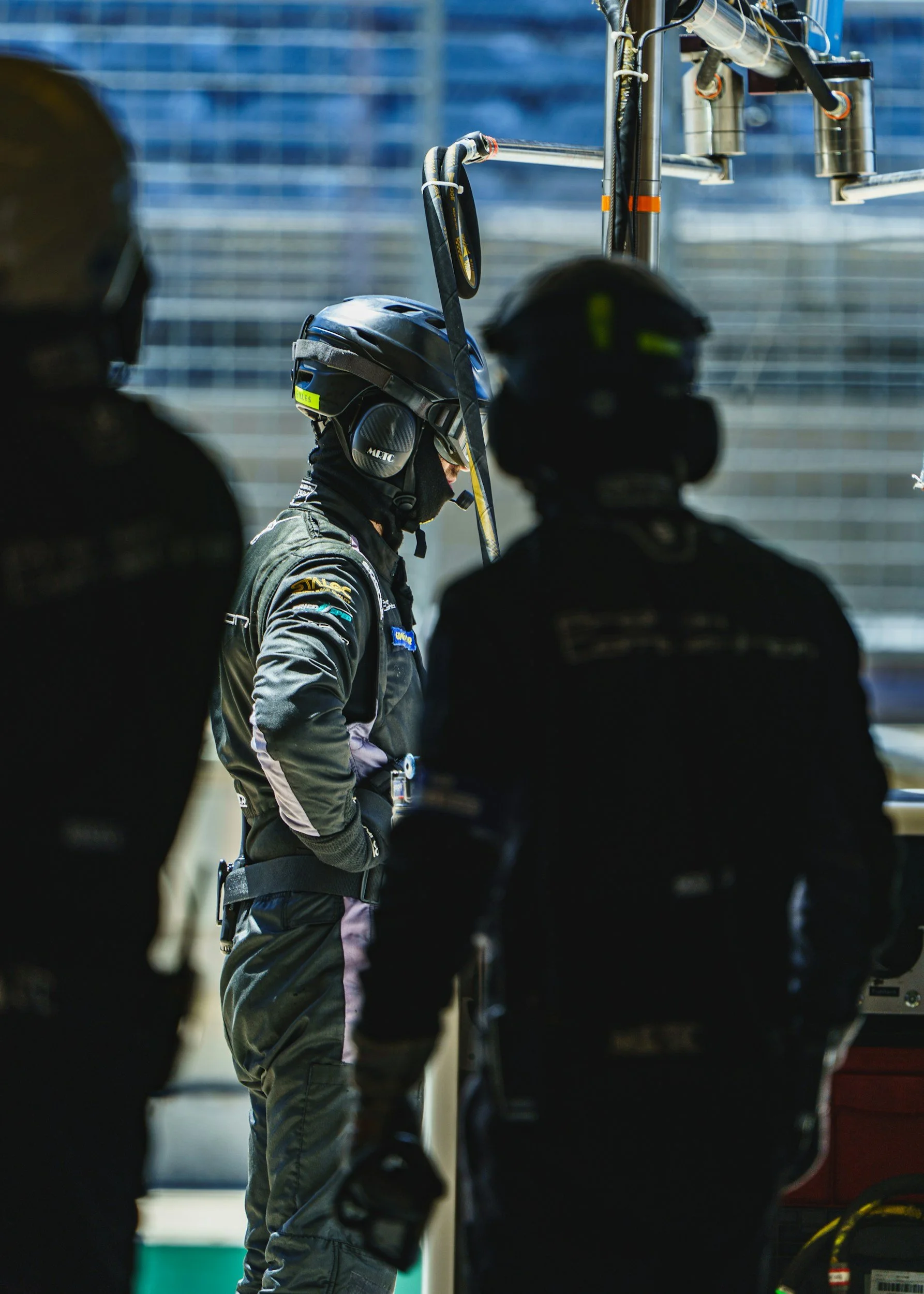 Race car driver standing in the garage with three crew members nearby, all wearing racing suits and helmets.