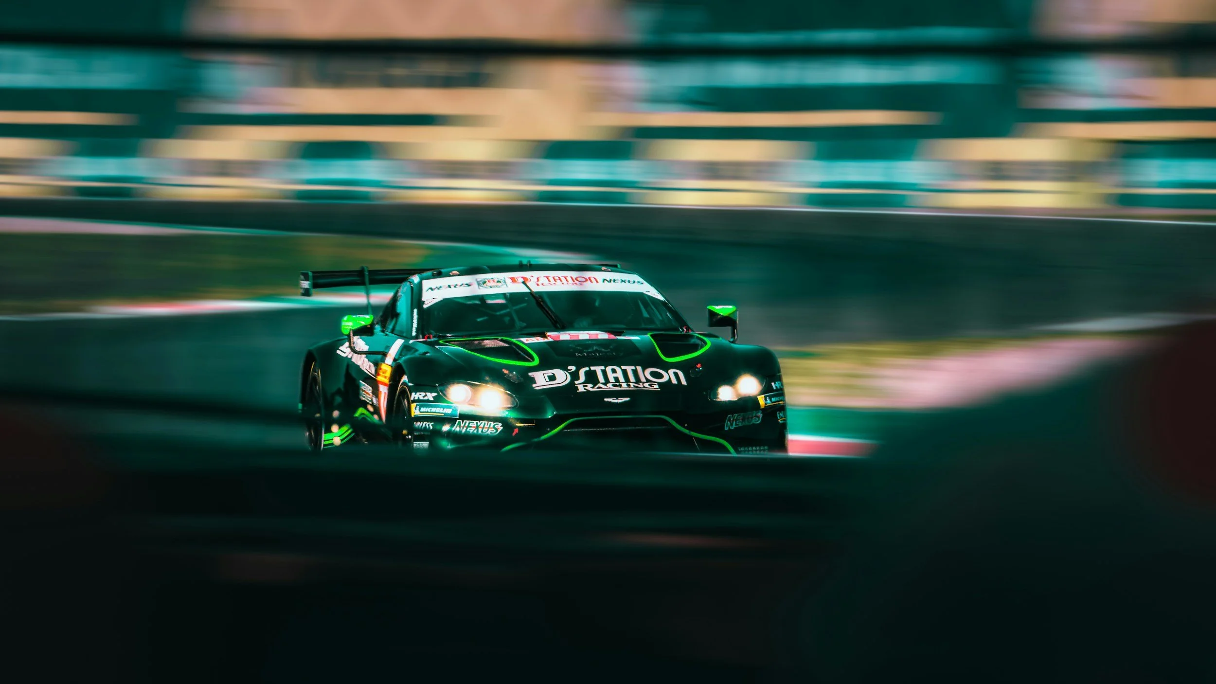 A race car on a track at night with blurred background, black with green accents, displaying 'D'STATION RACING' and various sponsor logos.