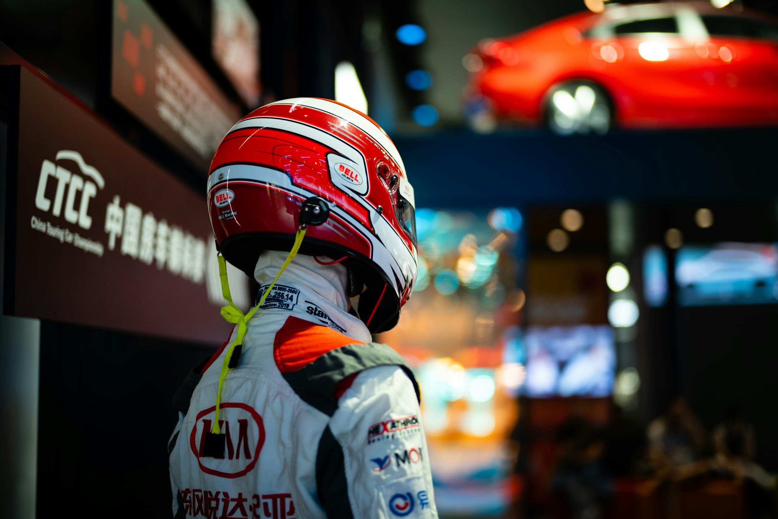 Race car driver wearing a red and white helmet and racing suit, standing in front of a digital display at a racing event.