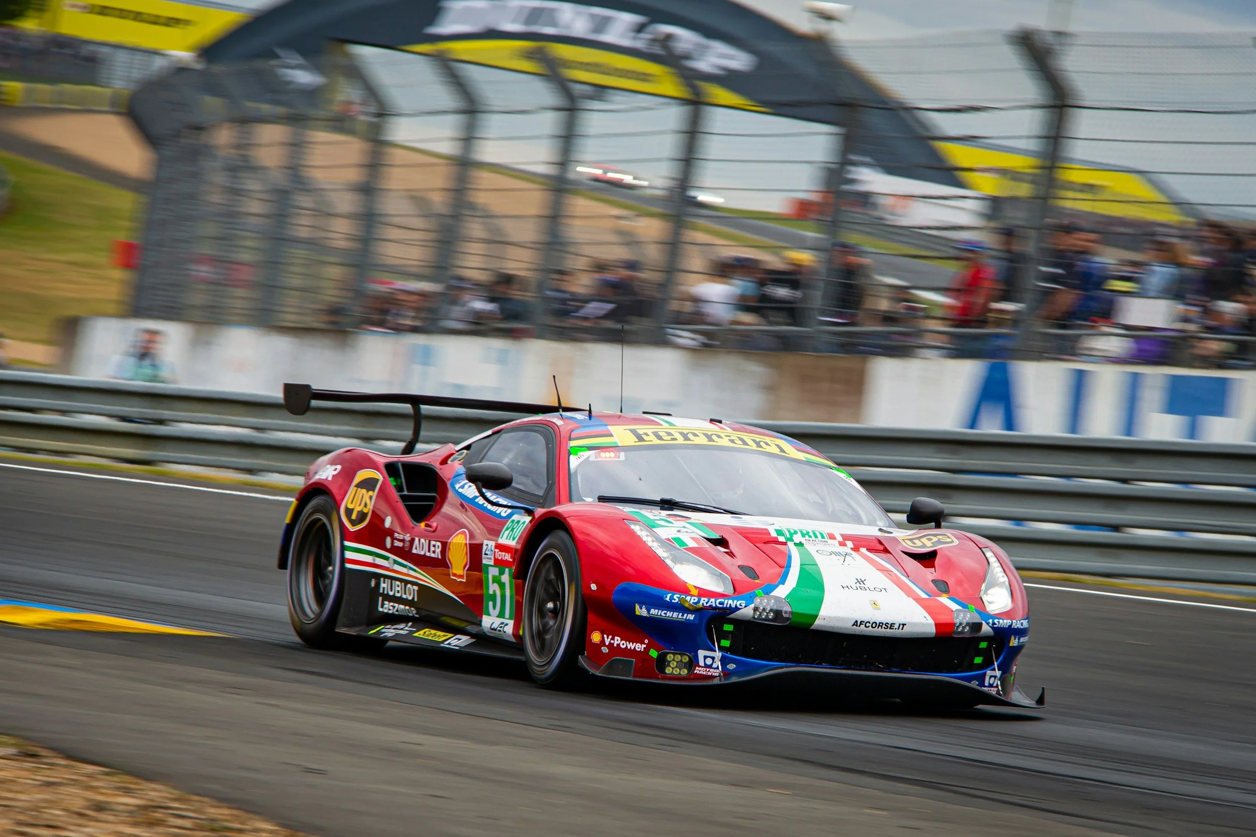 A red Ferrari race car with Lamborghini and other sponsor logos speeds on a racetrack with spectators and fencing in the background.