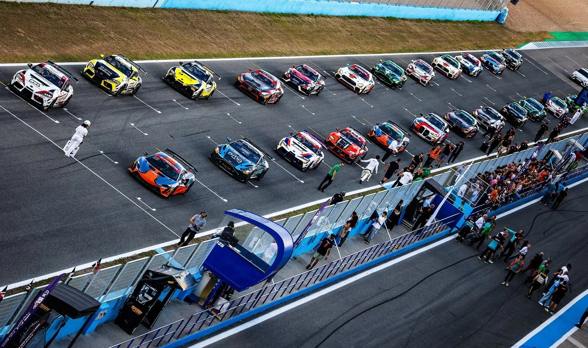 Race cars parked in the pit lane of a racetrack, with team members and spectators walking along the area.