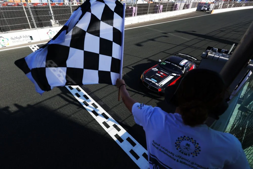 A person holding a checkered flag at a race track, with a race car crossing the finish line.