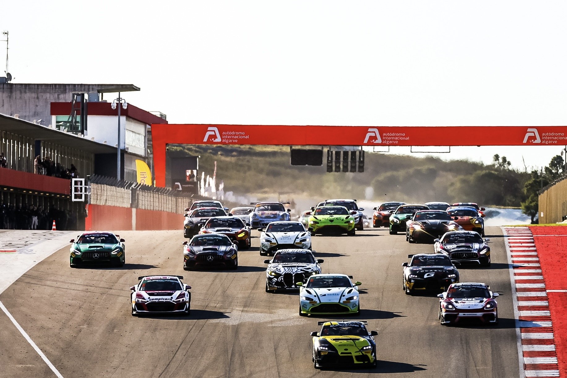 Multiple race cars on the track at Autódromo Internacional Algarve, with some cars leading near the camera and more cars in the background, approaching the finish line under a red bridge.