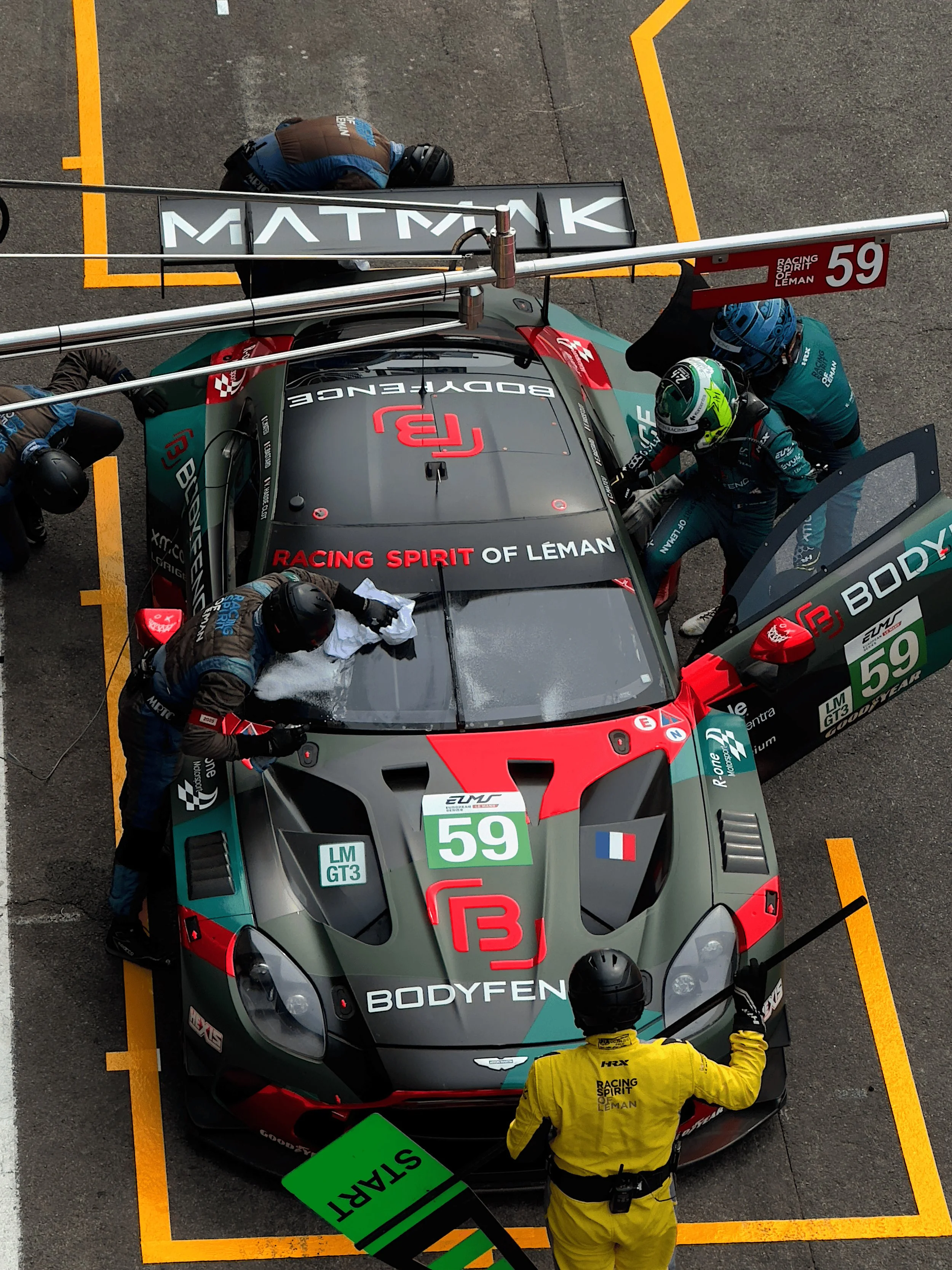 Race car in pit stop with crew members fueling and maintaining it, overseen by a marshal in yellow.