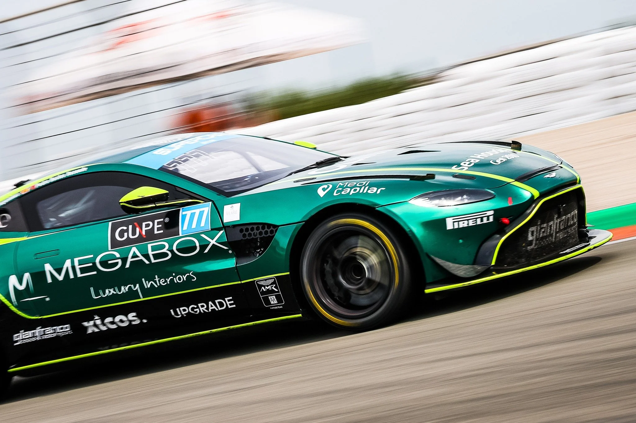 Green race car with sponsorship logos, moving quickly on a race track with blurred background.