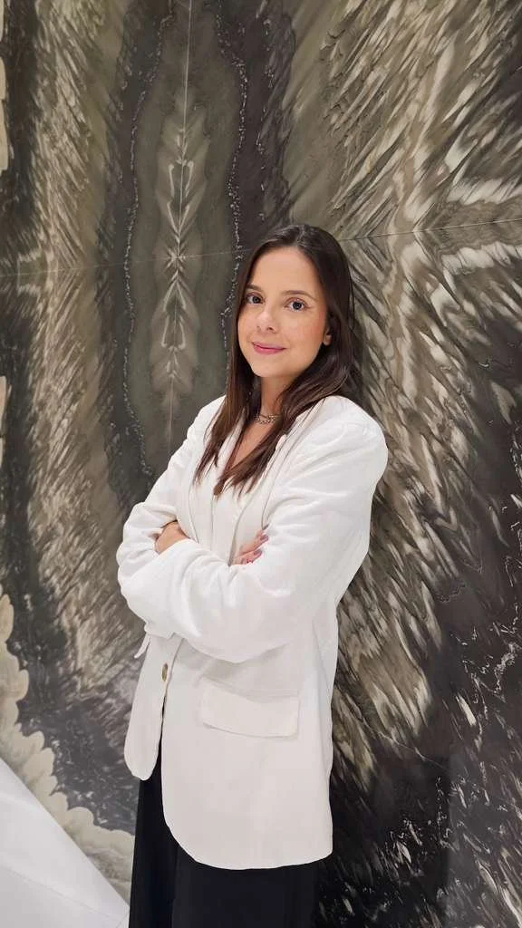 Yasmin Furtado an interior designer with long dark hair wearing a white blazer and black pants, standing with her arms crossed in front of a large marble-patterned wall during Coverings Show.