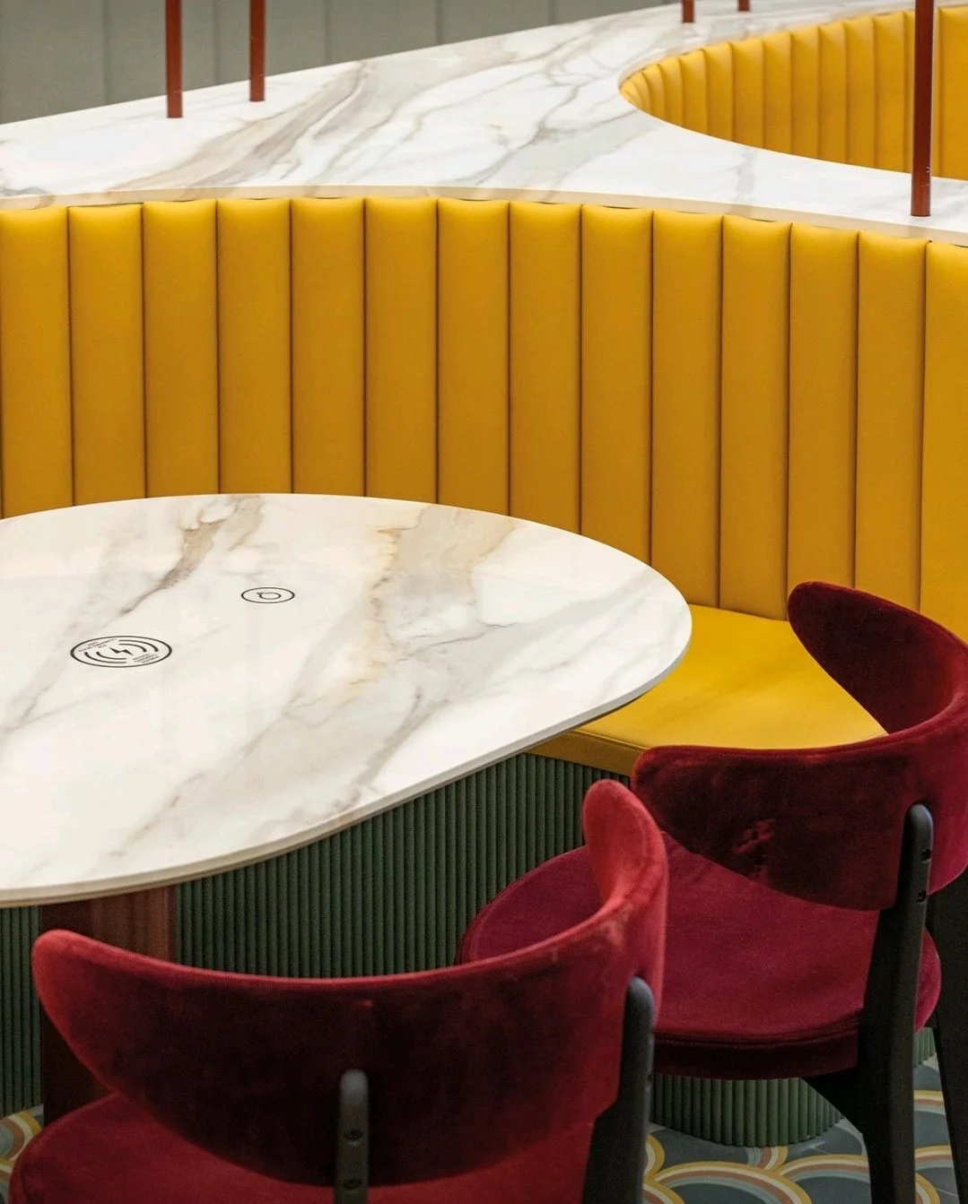 Modern restaurant booth featuring a custom-shaped porcelain table with built-in hidden phone charging, a yellow channeled back banquette, and velvet burgundy chairs.
