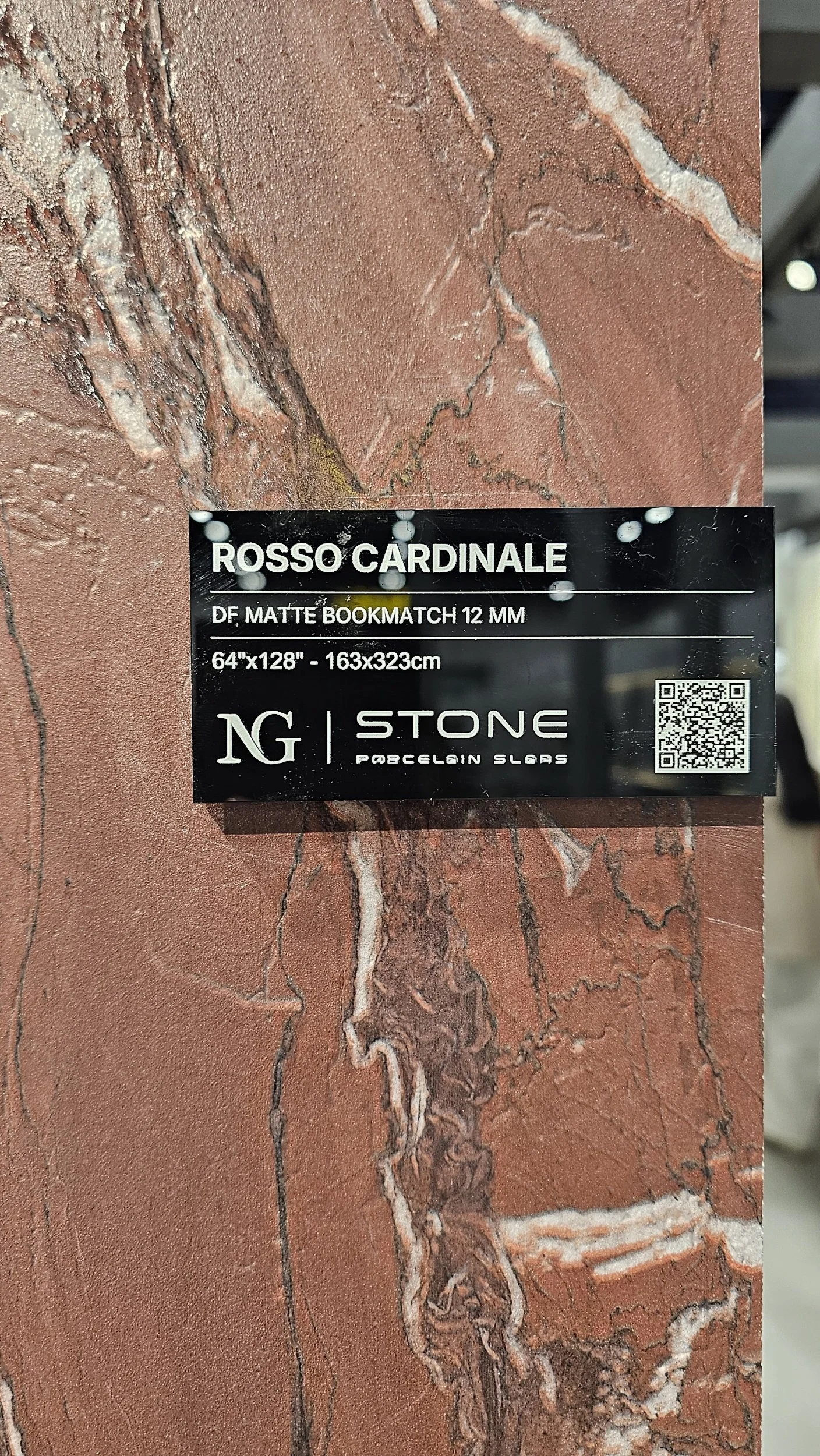 A close-up of a Rosso Cardinale porcelain slab by NG Stone featuring a matte bookmatch finish, with a black specification plaque detailing its 12mm thickness and $64" \times 128"$ dimensions