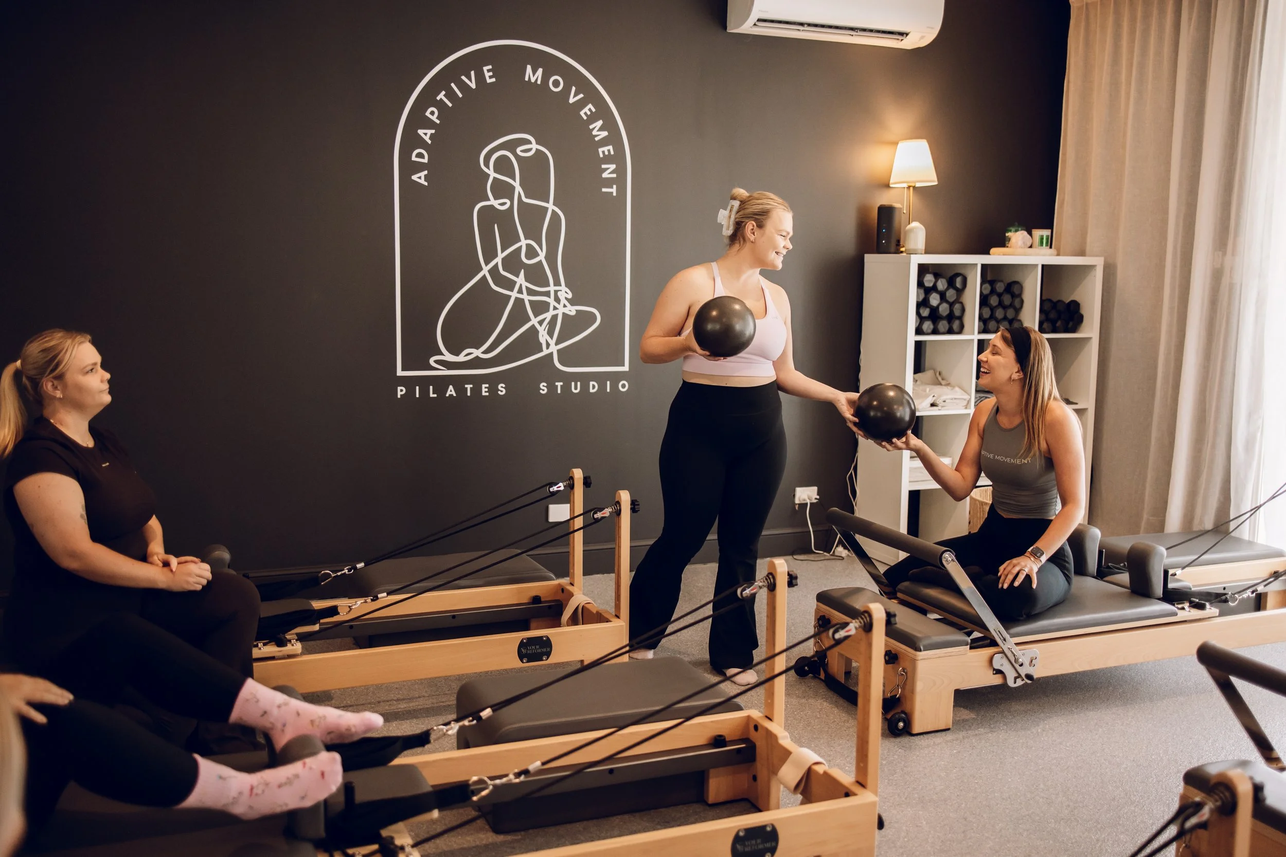 Women participating in a Pilates class using resistance bands while seated on Pilates reformers in a fitness studio with black and wooden interior decor.