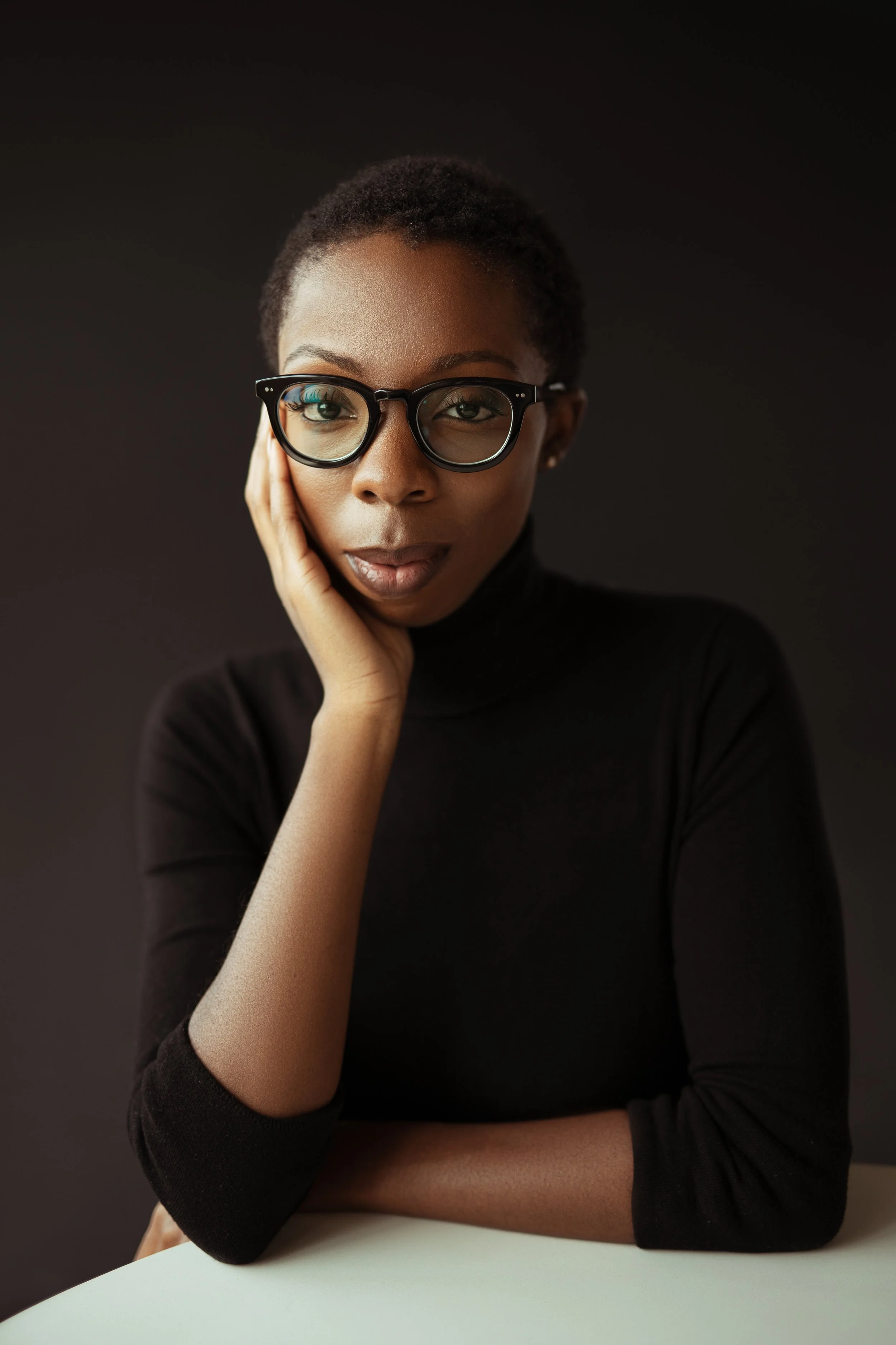 A young woman with dark skin wearing glasses and a black turtleneck, resting her face on her hand, sitting at a white table against a dark background.