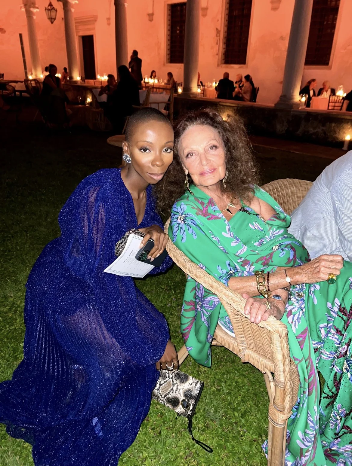 Two women posing for a photo at a nighttime outdoor event. One woman is wearing a shiny blue evening gown, and the other is wearing a green floral dress. They are sitting and kneeling on the grass, with a patio, tables, and people in the background.
