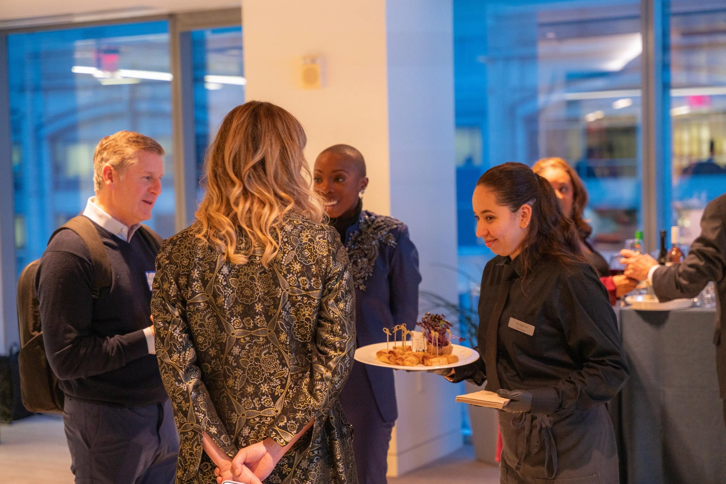 A group of people at a social gathering, talking to a server holding a plate with snacks, in a modern indoor setting.