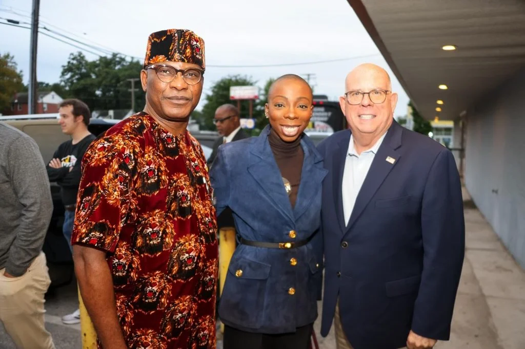 Three people standing outdoors, smiling at the camera. The person on the left is wearing a colorful, patterned shirt and a matching hat. The woman in the middle is wearing a dark blue blazer with gold buttons. The man on the right is dressed in a dar