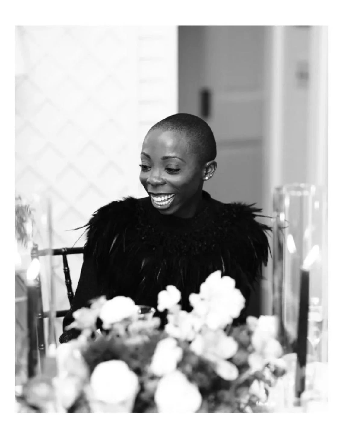 A smiling woman with a short buzz cut hairstyle, wearing a black top with feathered embellishments, sitting at a table with a blurred floral centerpiece in the foreground.