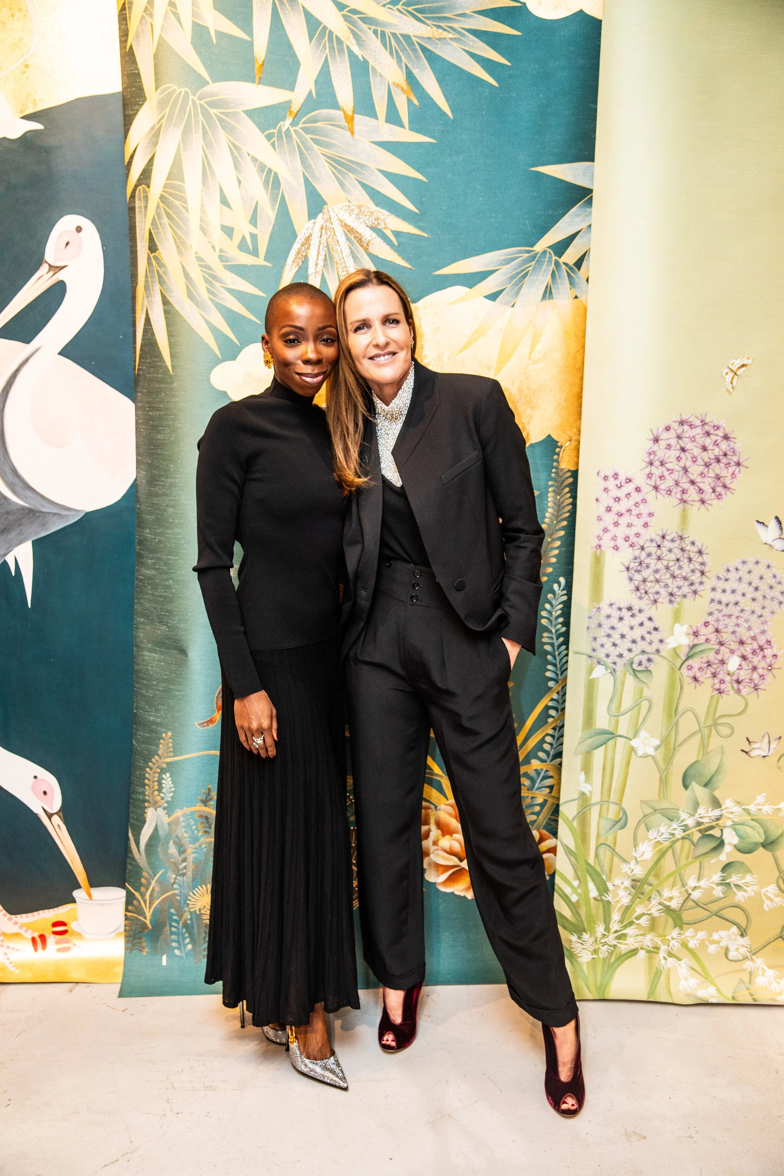 Two women in black outfits standing in front of a colorful nature-themed backdrop featuring storks, bamboo, flowers, leaves, and butterflies.