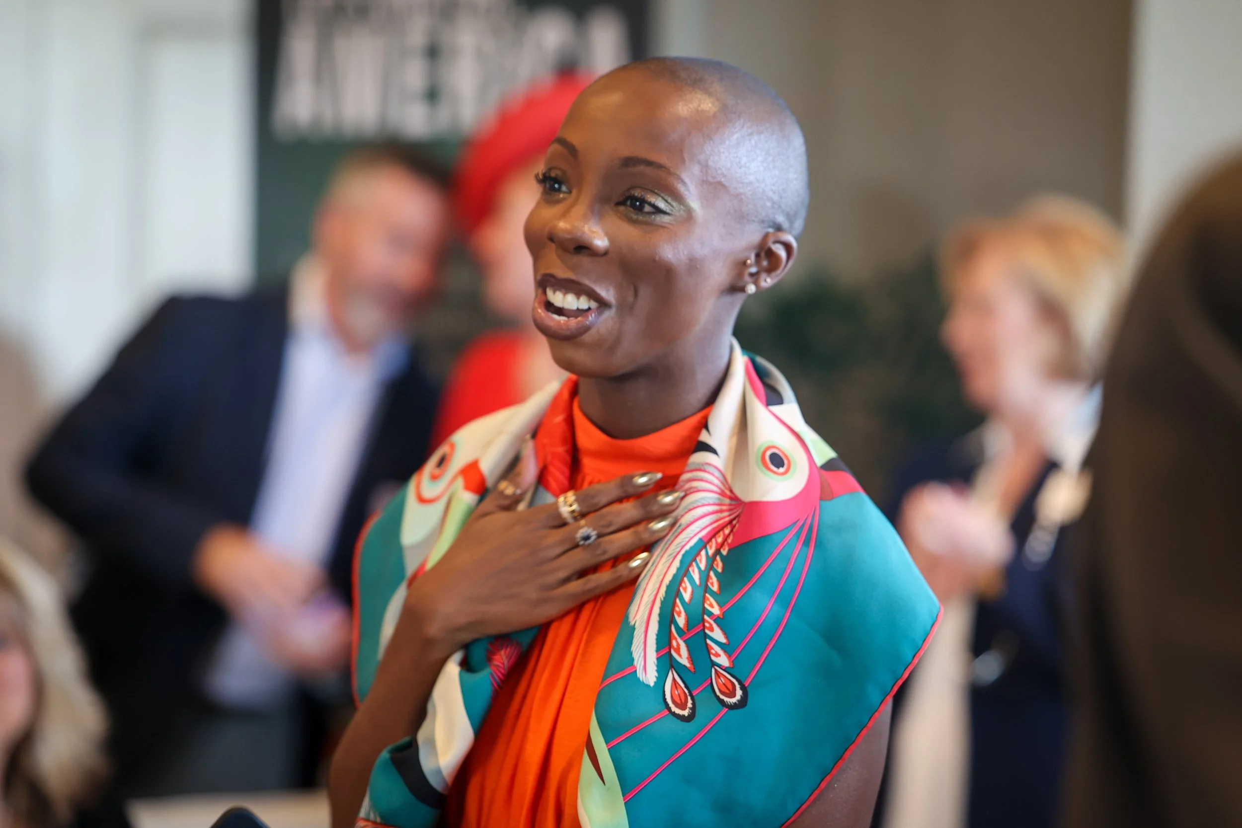 A woman with a shaved head, wearing a colorful scarf and orange top, smiling and touching her chest, at a social gathering with blurred people in the background.